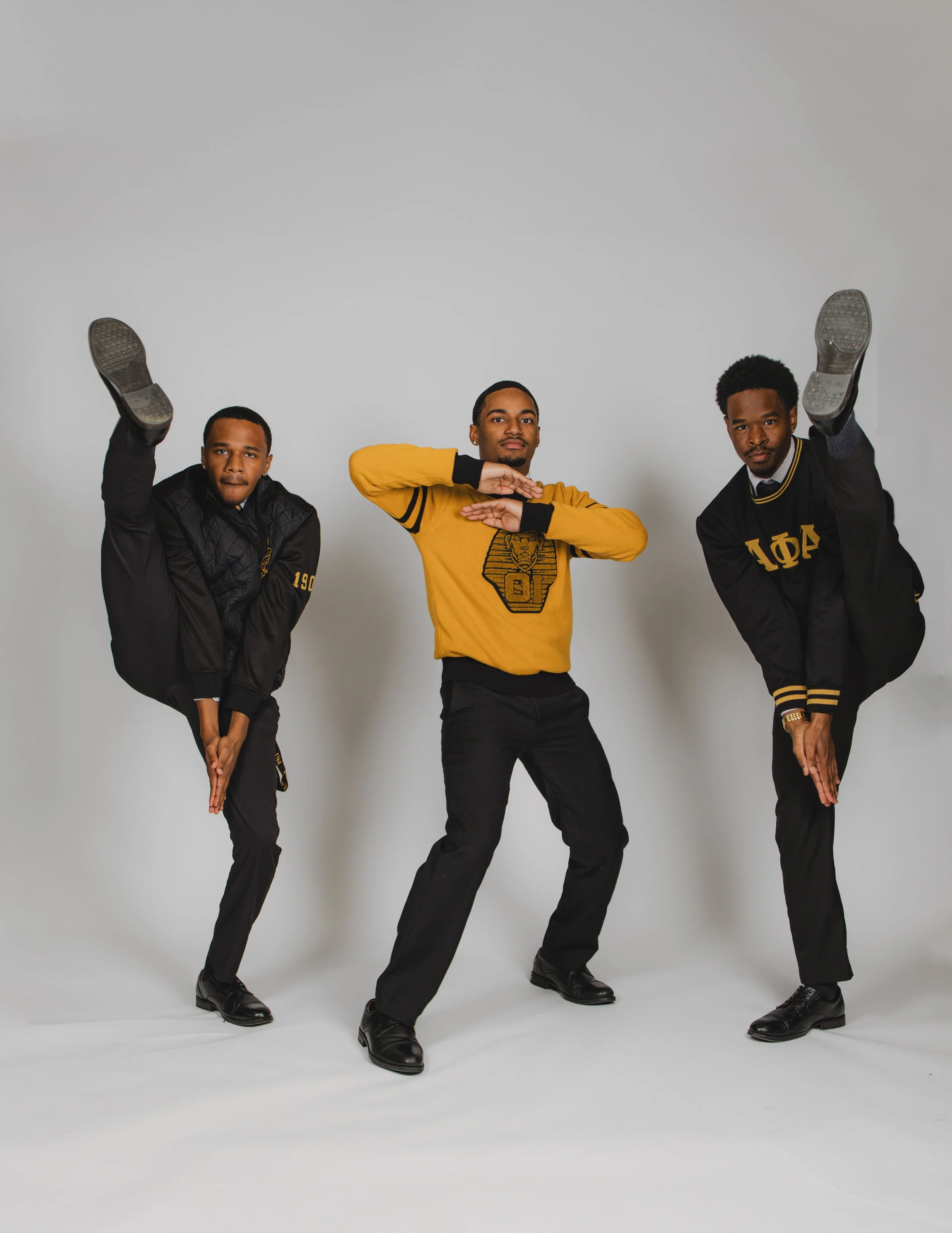 Three young men in black and yellow clothing in a studio, balancing on one leg with one hand reaching downward and the other arm bent across their chest.