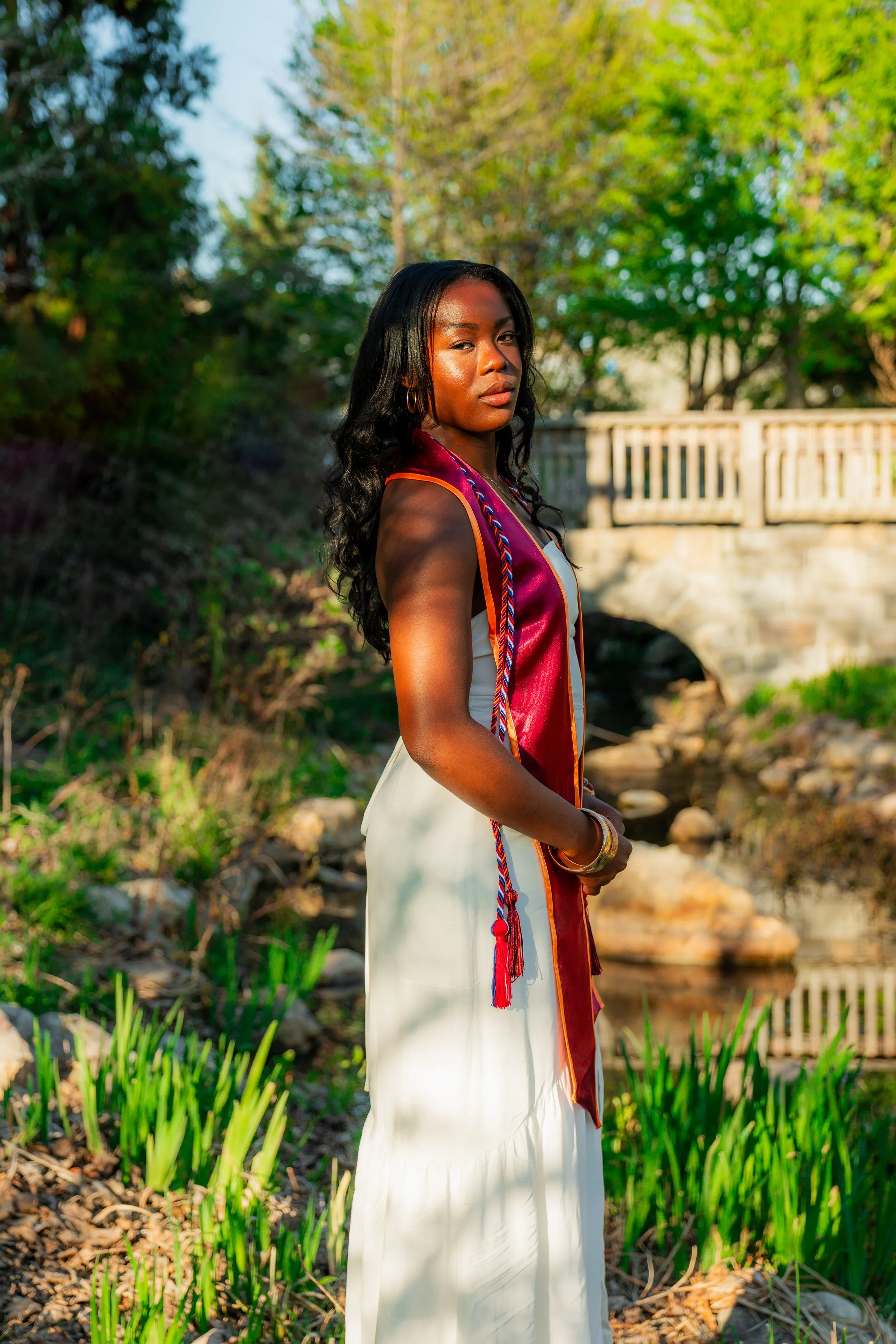 A young woman standing outdoors near a creek, wearing a white dress and a maroon sash with orange trim, with trees and a wooden bridge in the background, illuminated by warm sunlight.