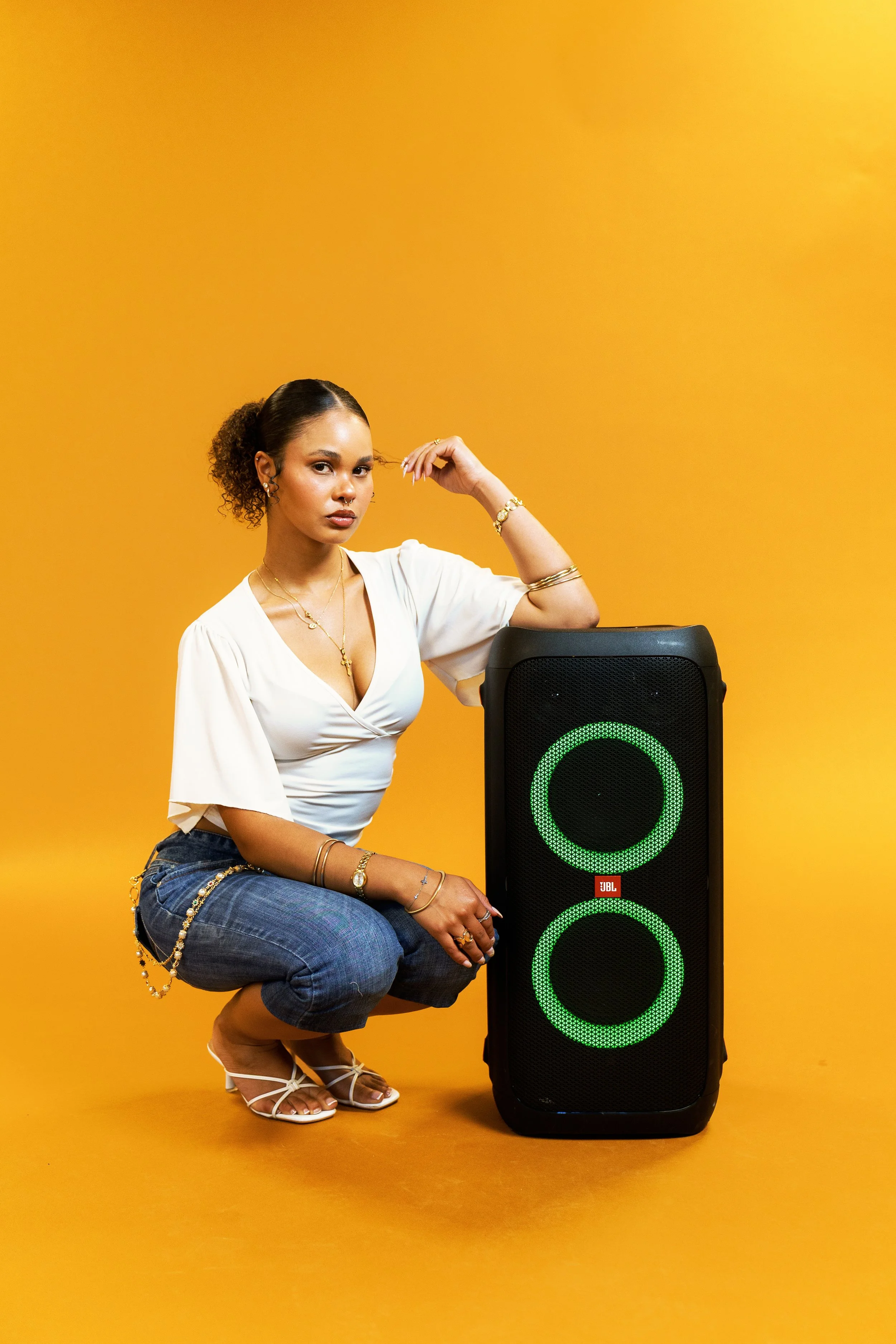 A woman with dark curly hair tied back, wearing a white top, blue jeans with jewelry, and white sandals, is squatting next to a large black speaker with green lights on a yellow background.