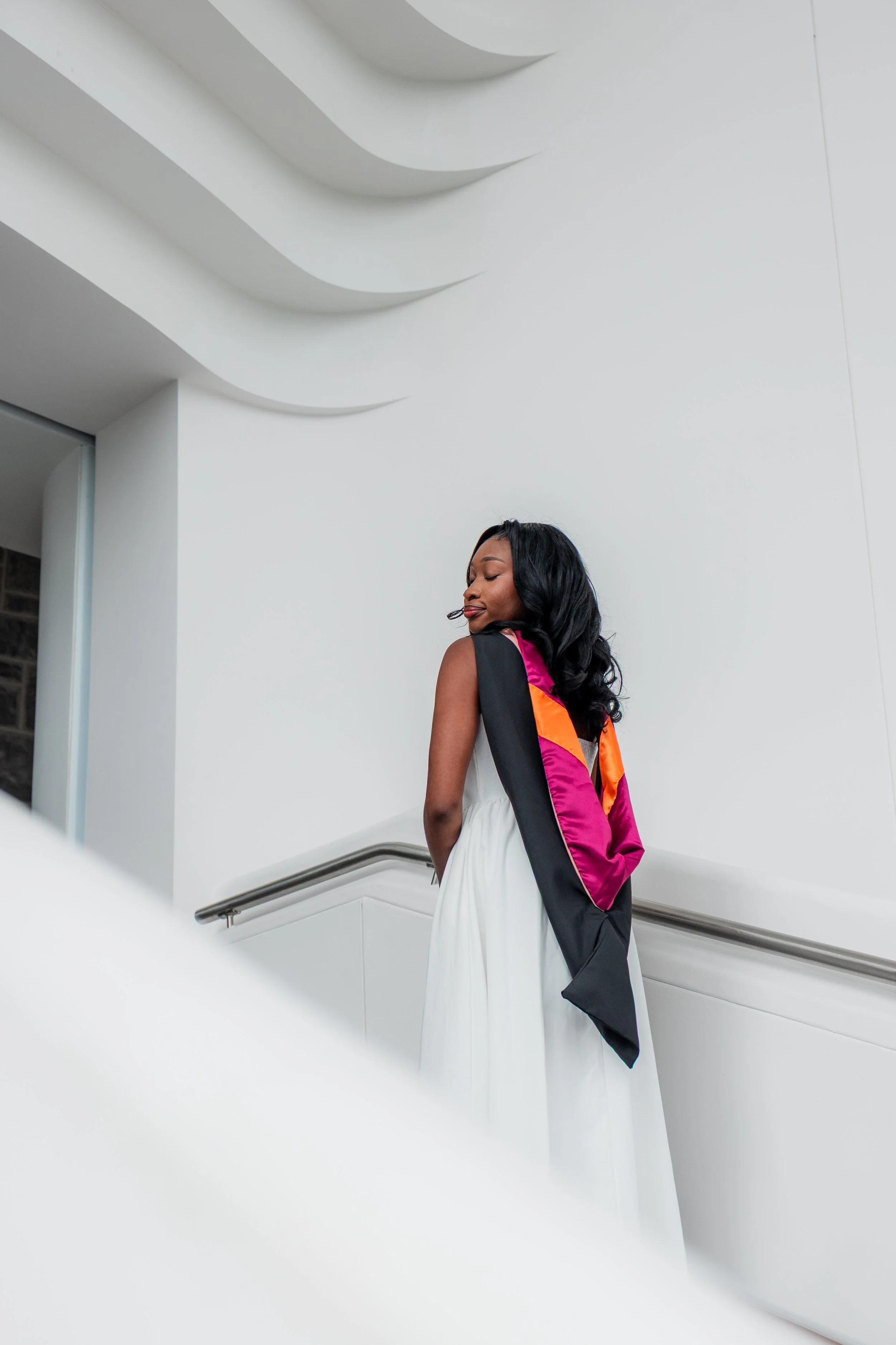 A woman with long black hair wearing a graduation gown and sash, standing indoors on a staircase with white walls and modern architecture.