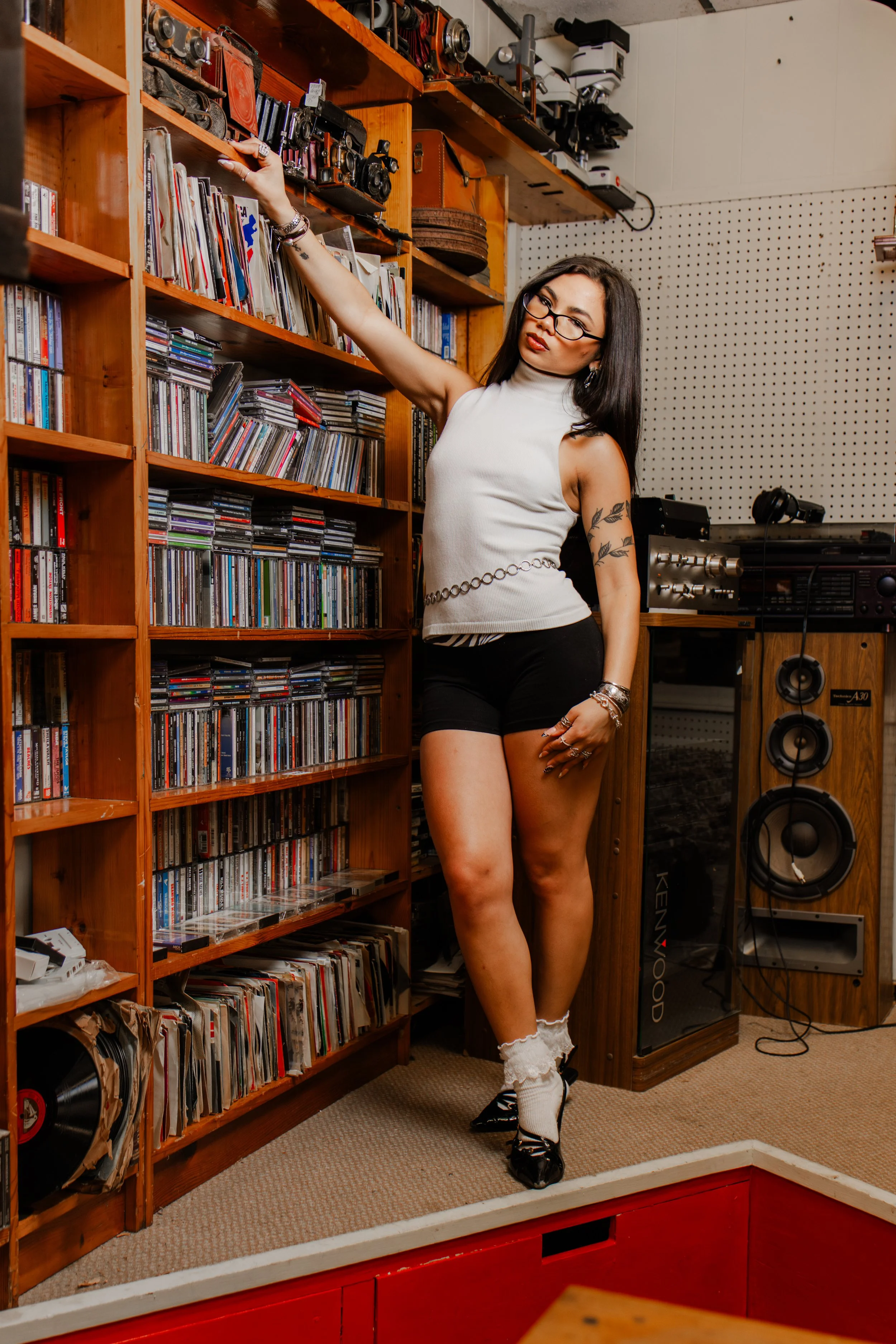 A woman with glasses and dark hair poses in a room with wood shelves filled with CDs, cameras, and audio equipment. She wears a sleeveless white top, black shorts, and black shoes with white socks. She is standing on a small platform in front of a pe
