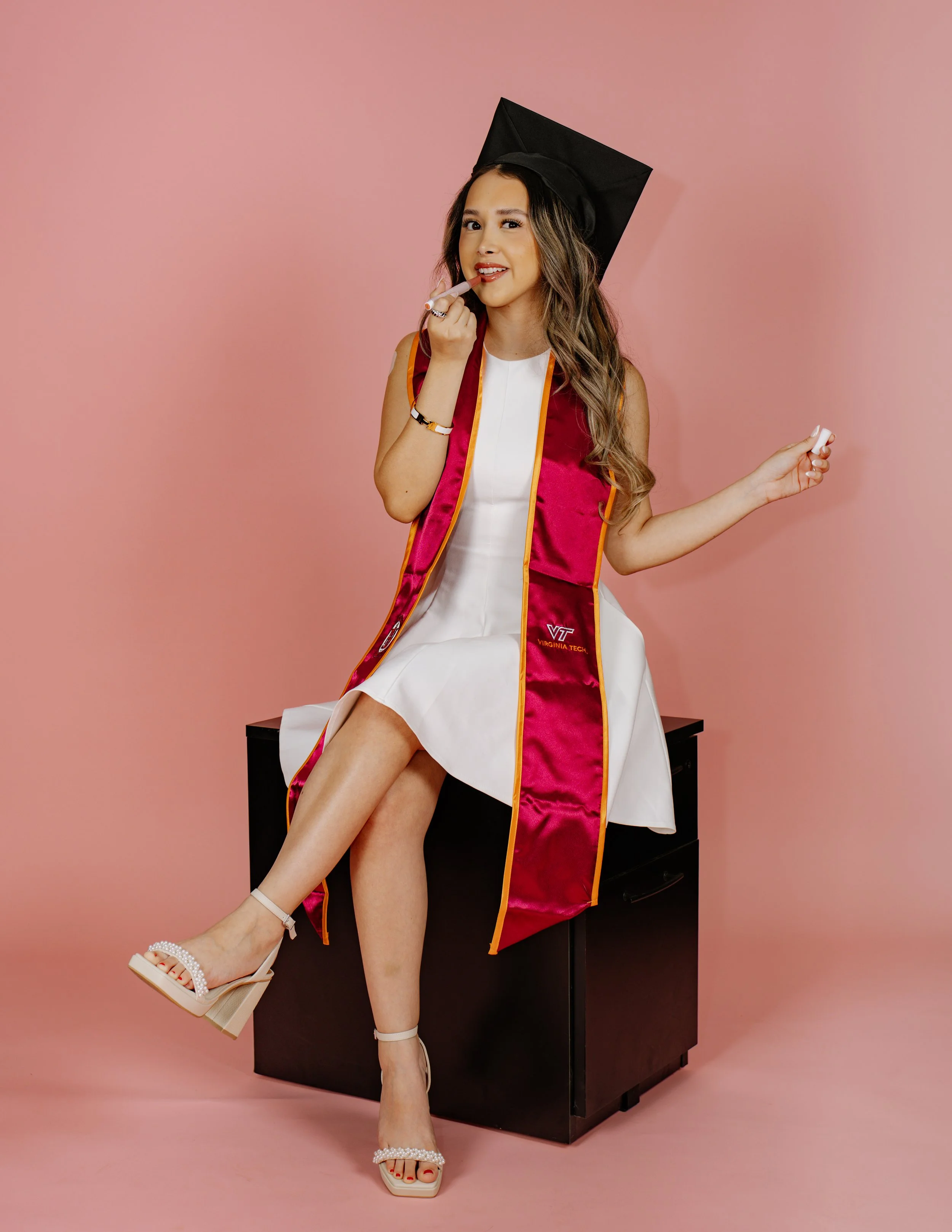 A young woman in a white dress and heels, wearing a graduation cap and gown, sitting on a black desk against a pink background, smiling and holding a diploma.