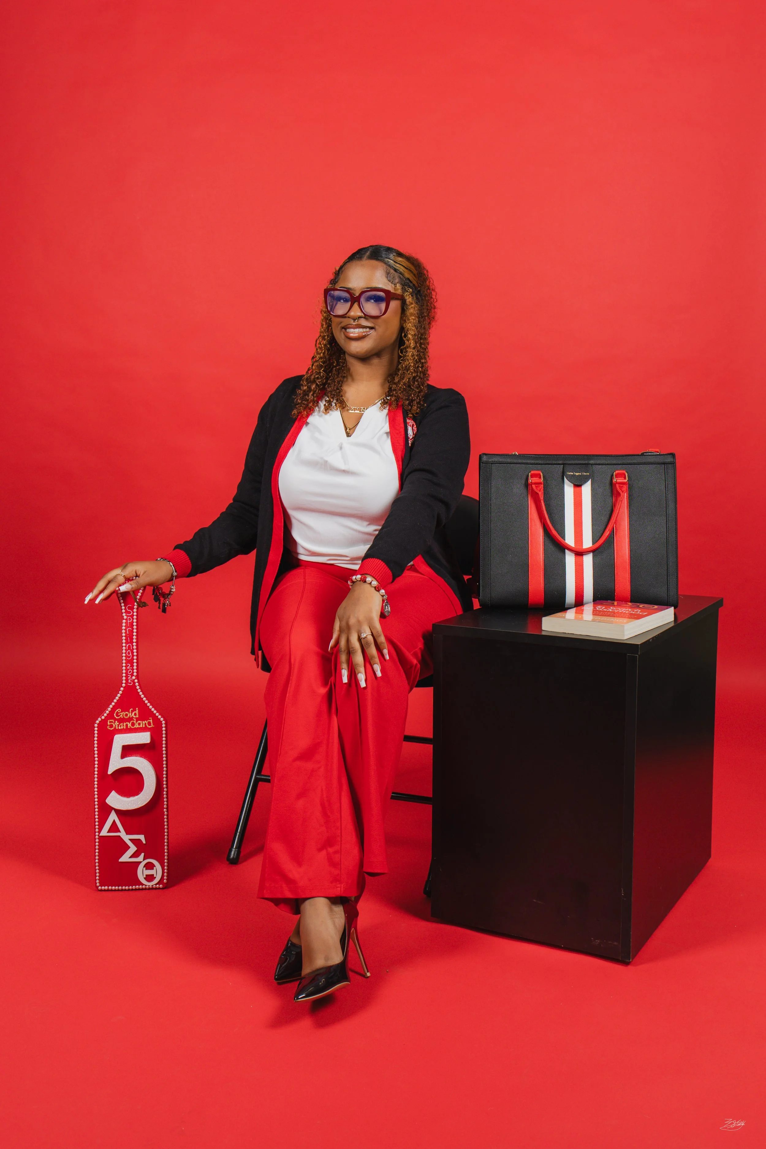 A woman with curly hair and glasses sitting on a black chair against a red background. She is wearing a black and red jacket, white shirt, and red pants, with black high heels. Beside her, on the floor, is a red and white sign with the number 5 and G