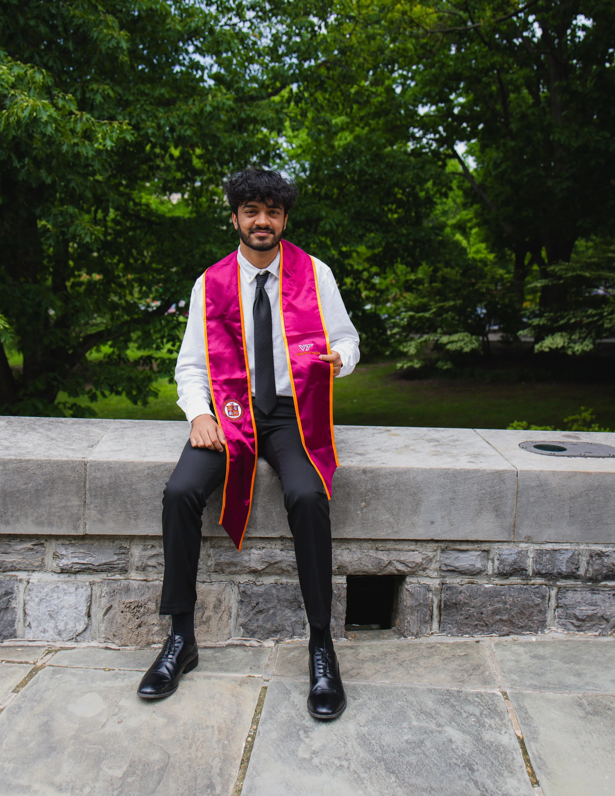 A young man in a graduation gown sitting on a stone ledge outdoors with greenery in the background.
