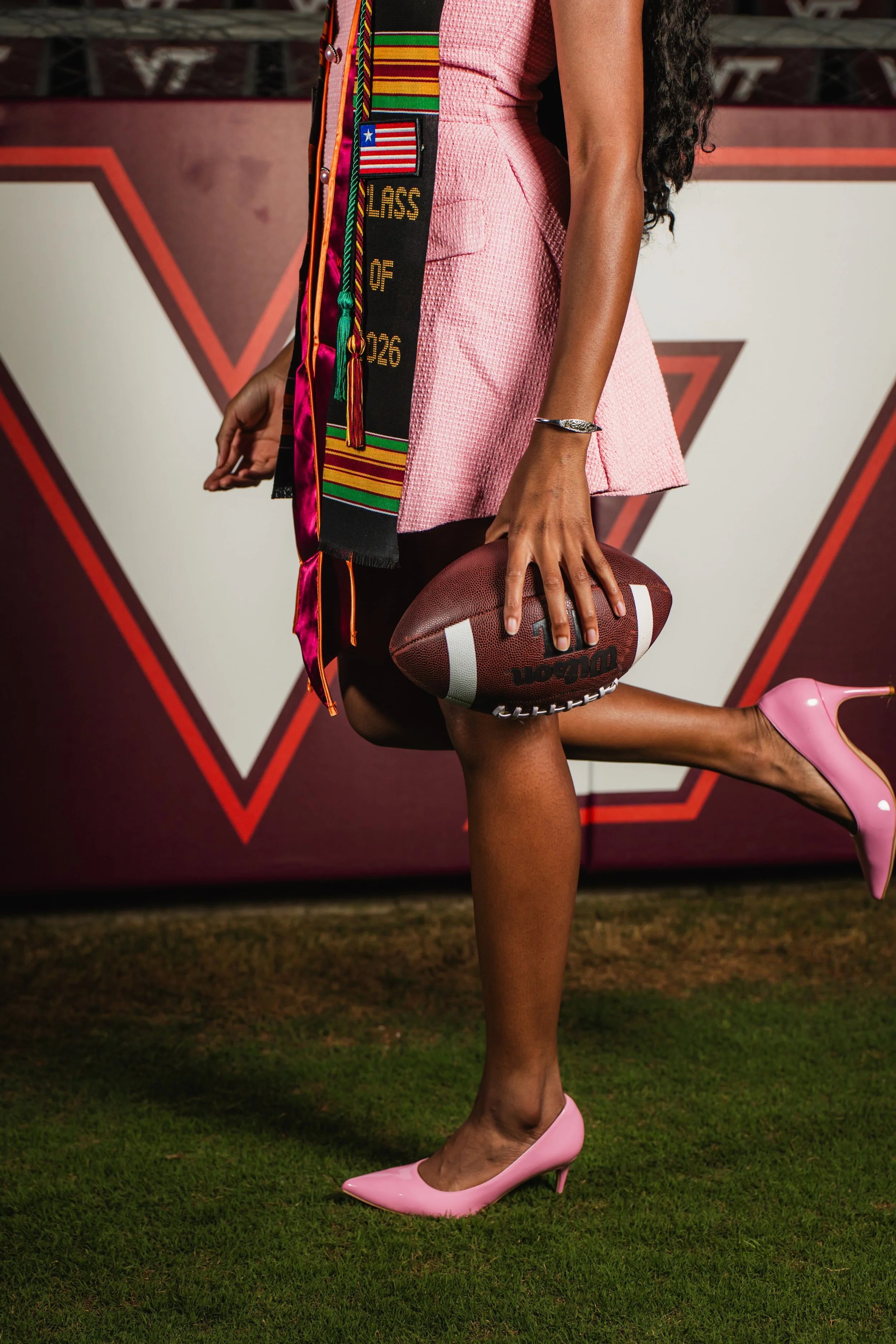 A woman in a pink textured dress with bold jewelry holds an American football while wearing pink high heels, standing on grass against a backdrop with geometric red and white designs.