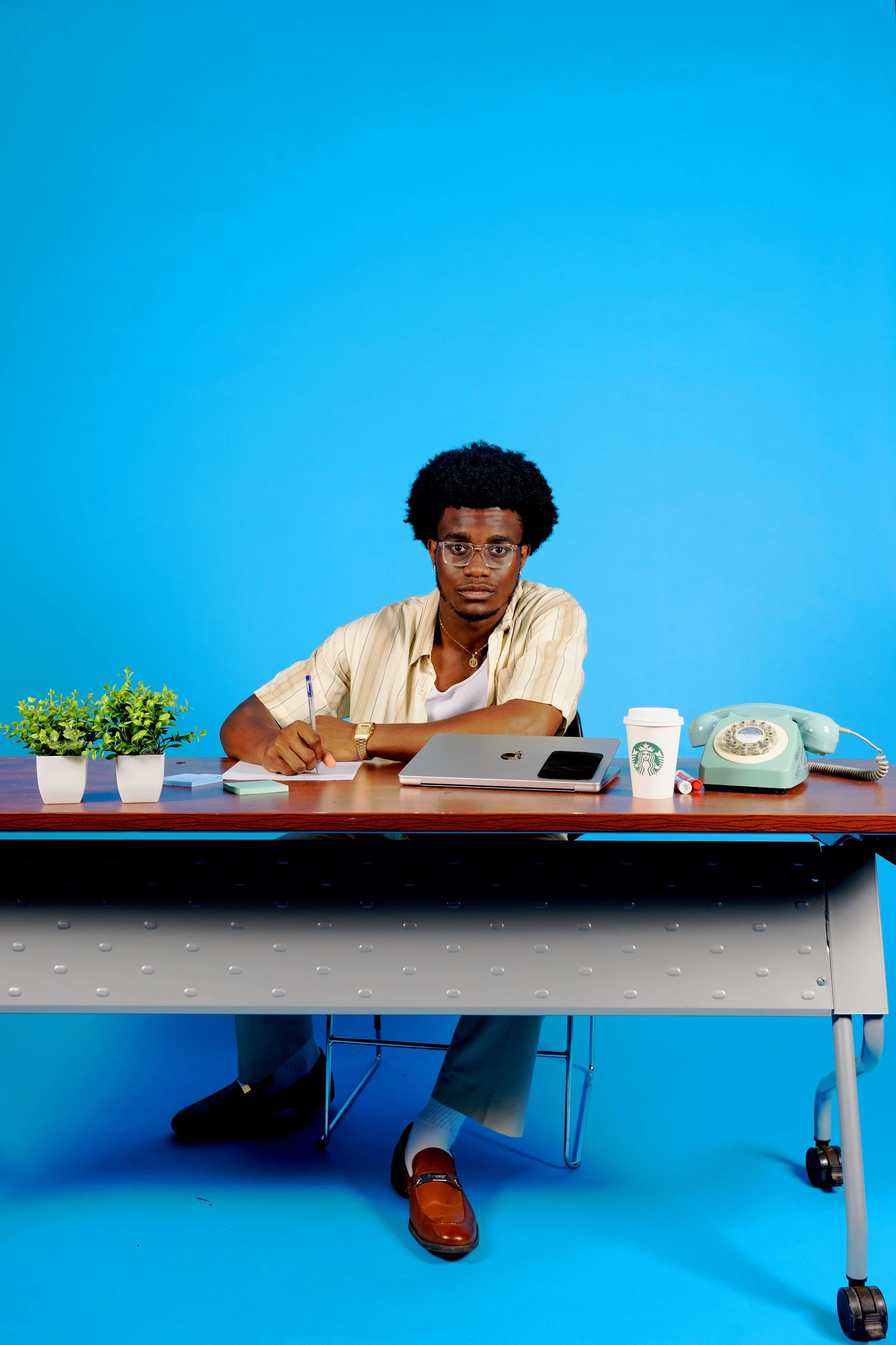 A man with glasses and a curly afro hairstyle sitting at a desk with a beige shirt, writing with a pen in paper. On the desk are a potted plant, Starbucks coffee cup, smartphone, laptop, and a light green rotary phone. The background is solid bright 