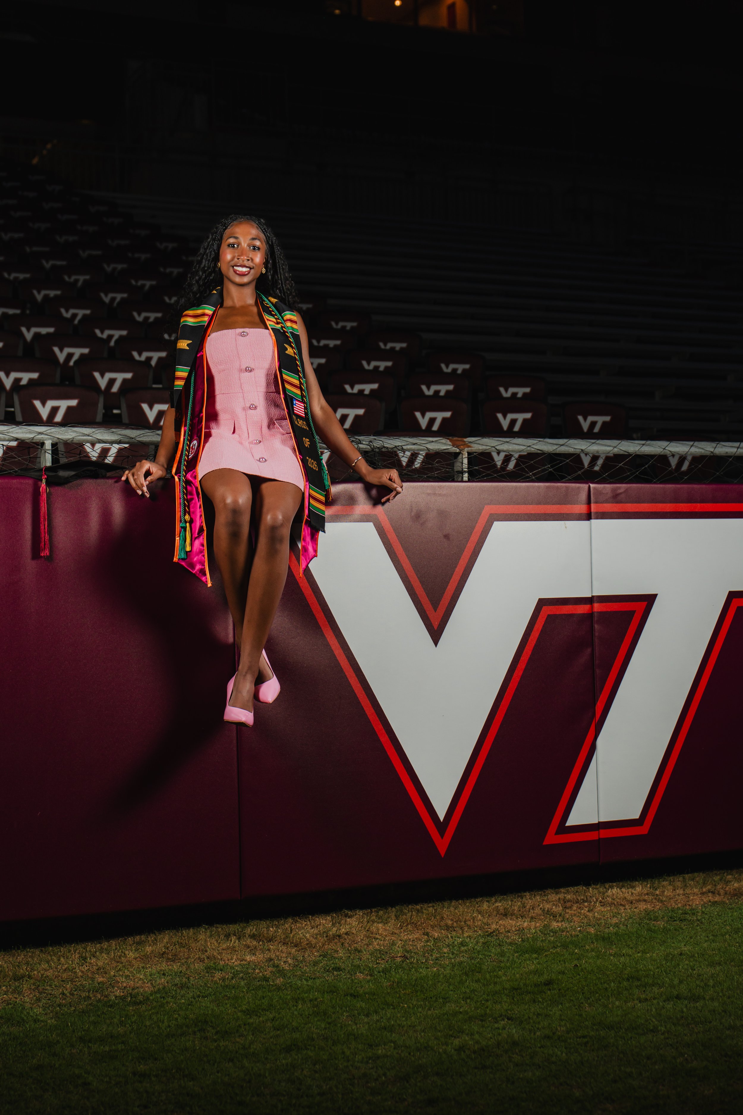 A woman in a pink dress with buttons, black cape with multicolored stripes, and pink high heels sitting on a maroon barrier at a sports stadium.