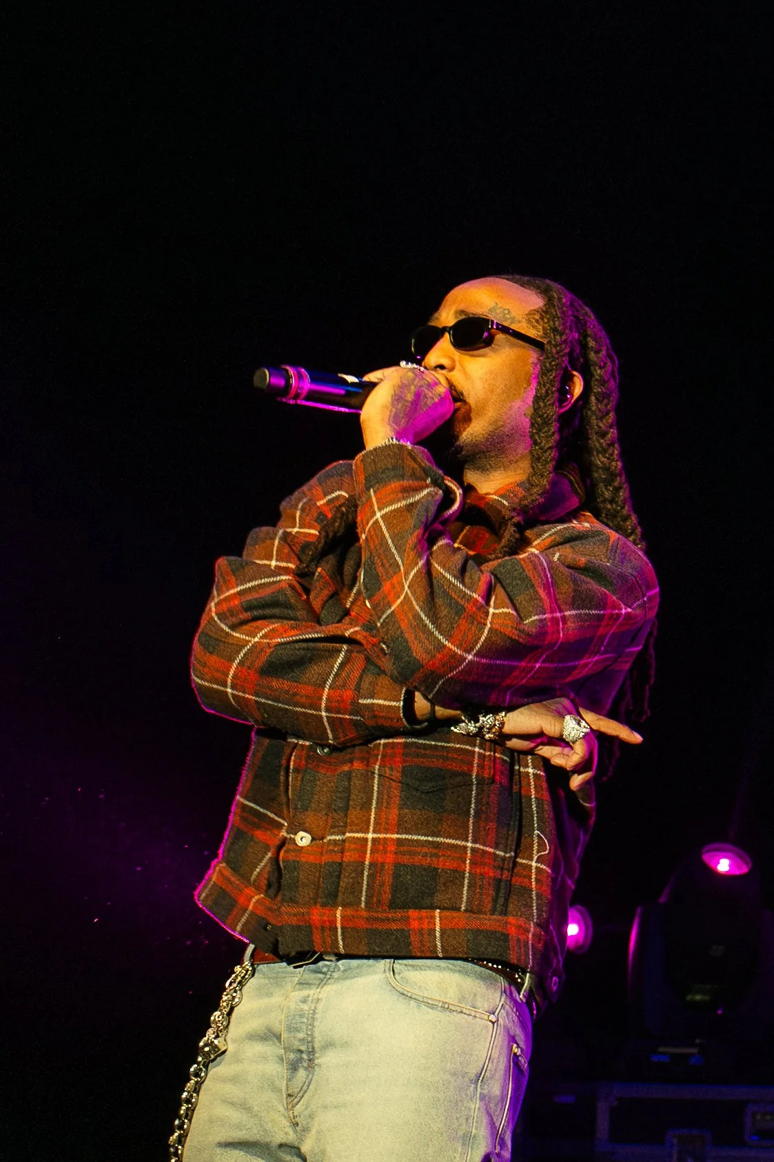 A male music performer called Quavo with long braided hair, wearing sunglasses, a plaid jacket, and light-colored pants, performing on stage with a microphone.