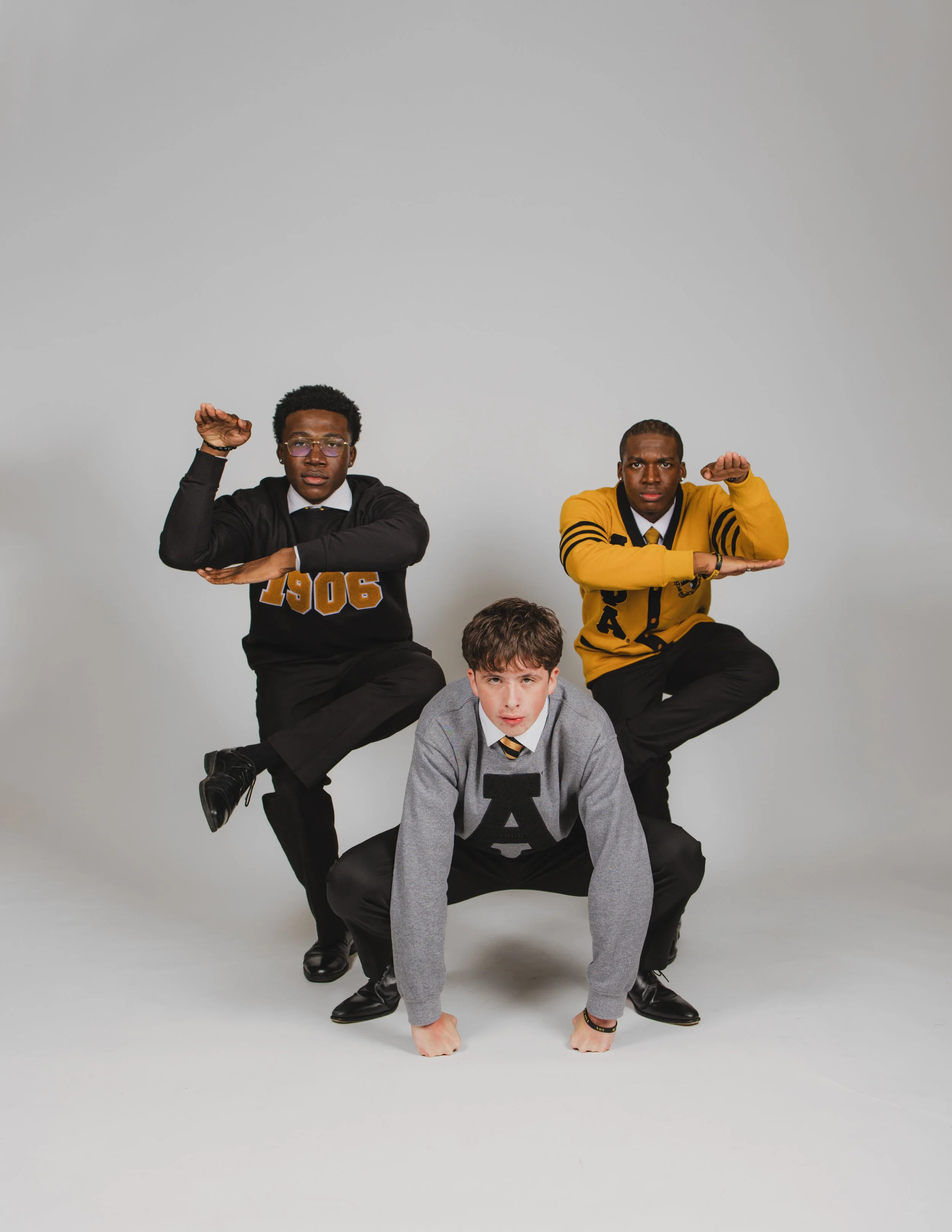 Three young men in casual and semi-formal clothing forming a human pyramid against a plain light gray background, with one person in a gray sweater at the bottom, and two others in black and yellow sweaters on top.