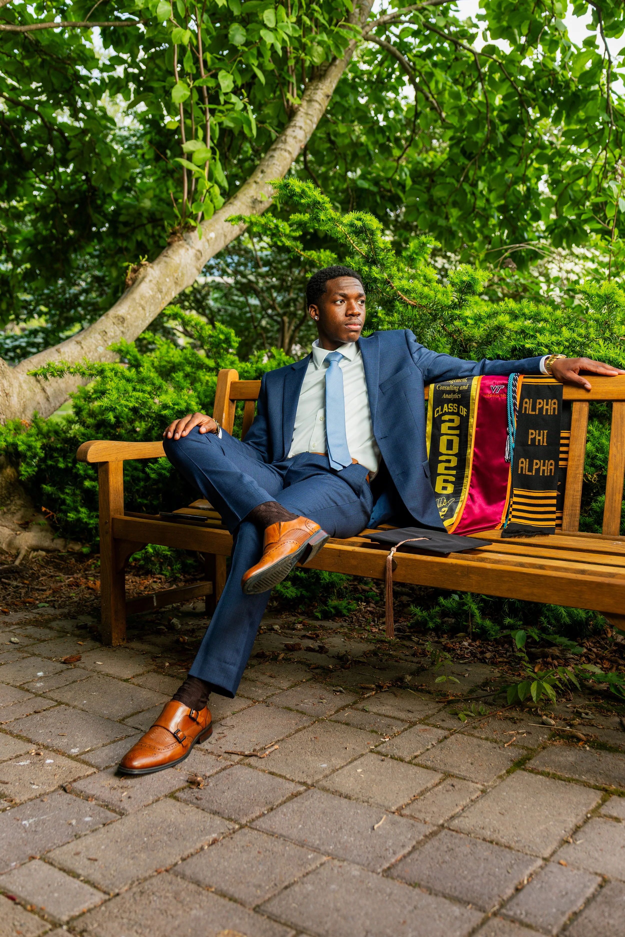 A young man in a blue suit sitting on a wooden bench in a park, surrounded by green trees and shrubs, with a graduation stole draped over the side of the bench.