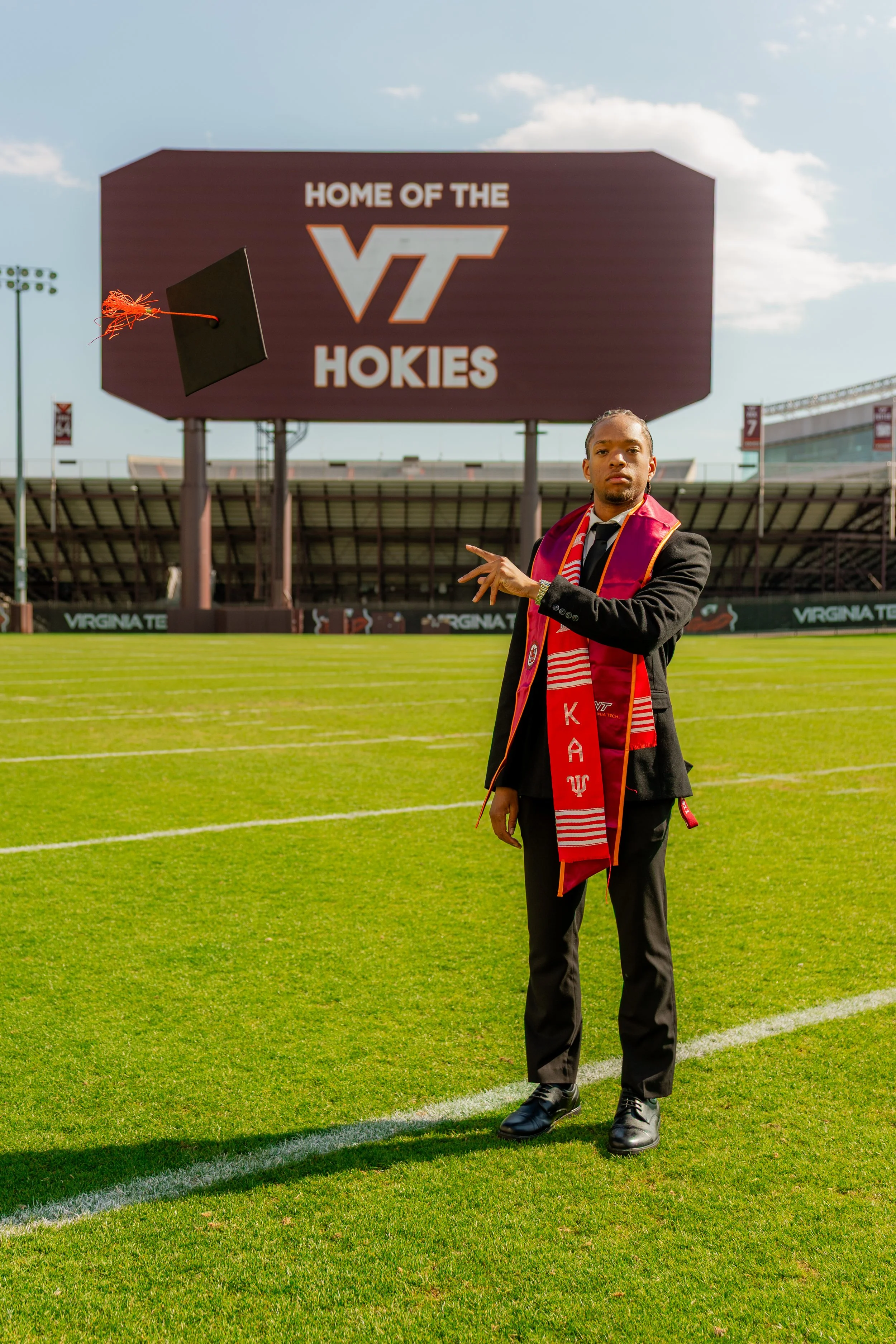 A young man in a suit and tie standing on a football field, wearing a red scarf with Greek letters, pointing at a sign that reads 'Home of the VT Hokies.' A black graduation cap is flying through the air.