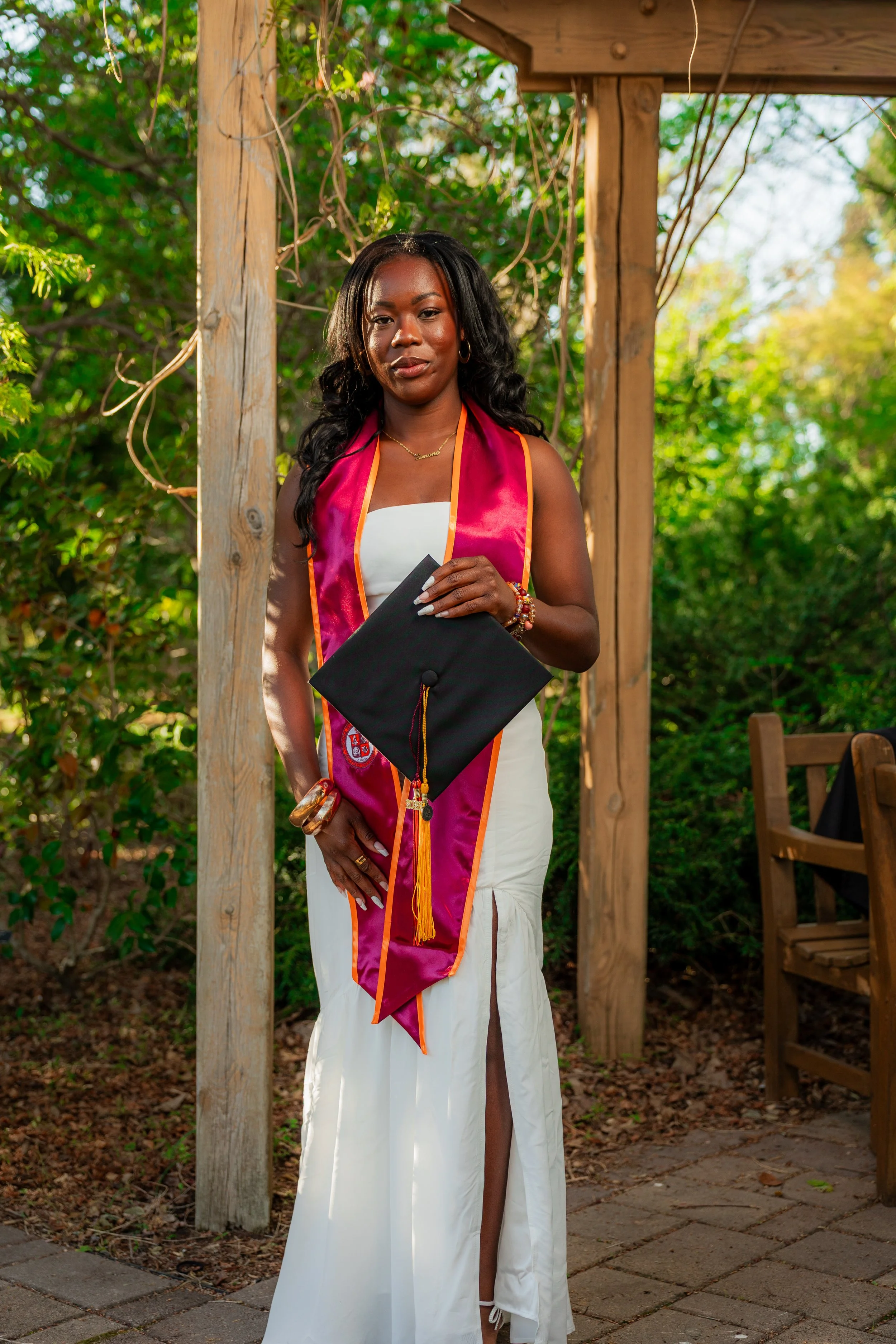 A woman in a white dress holding a graduation cap, standing outdoors next to a wooden post with greenery in the background.