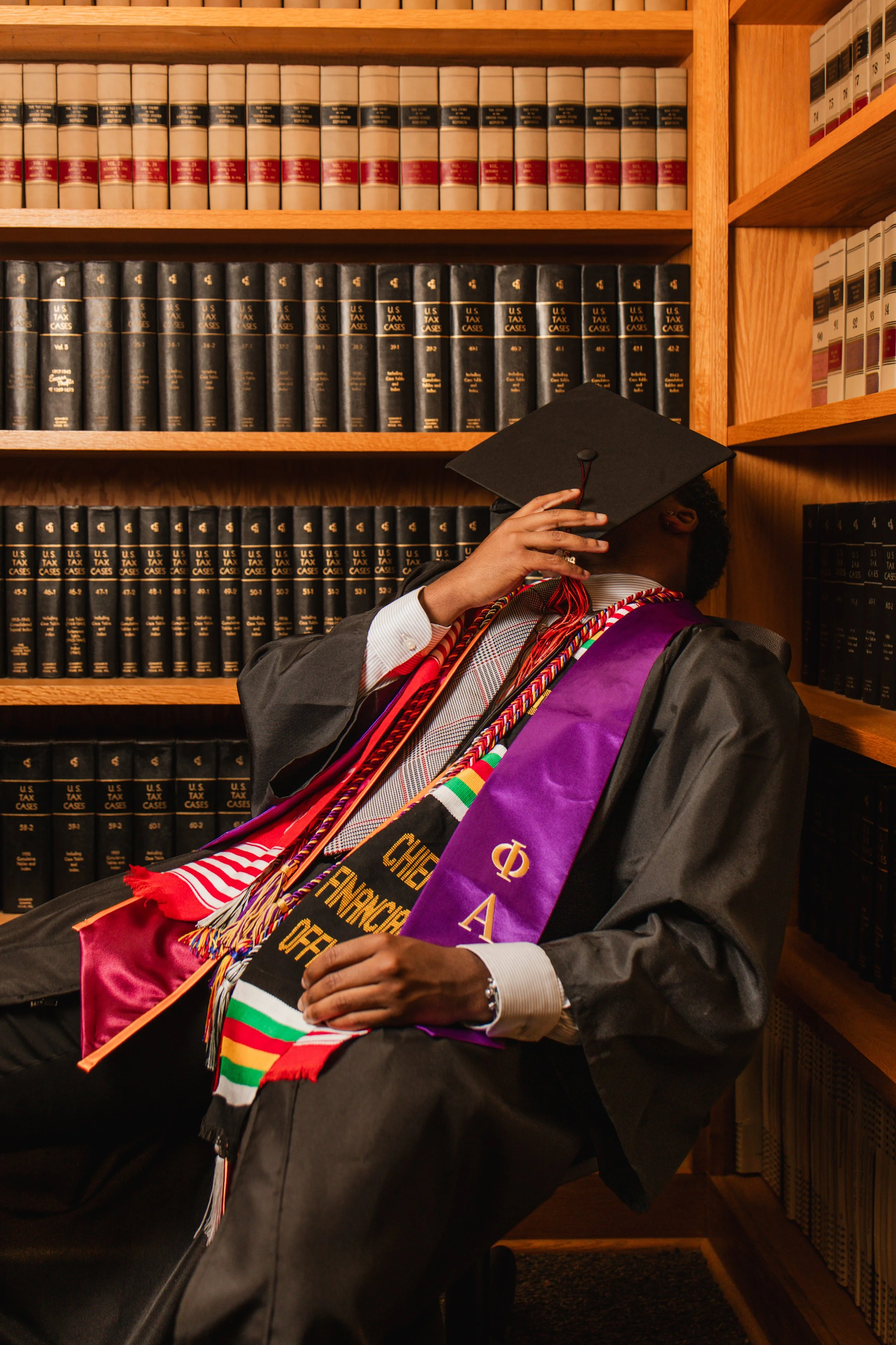 A person in academic regalia wearing multiple honor cords and stoles, sitting in front of a wooden bookshelf filled with law books and legal case reporters, partially covering their face with a graduation cap.