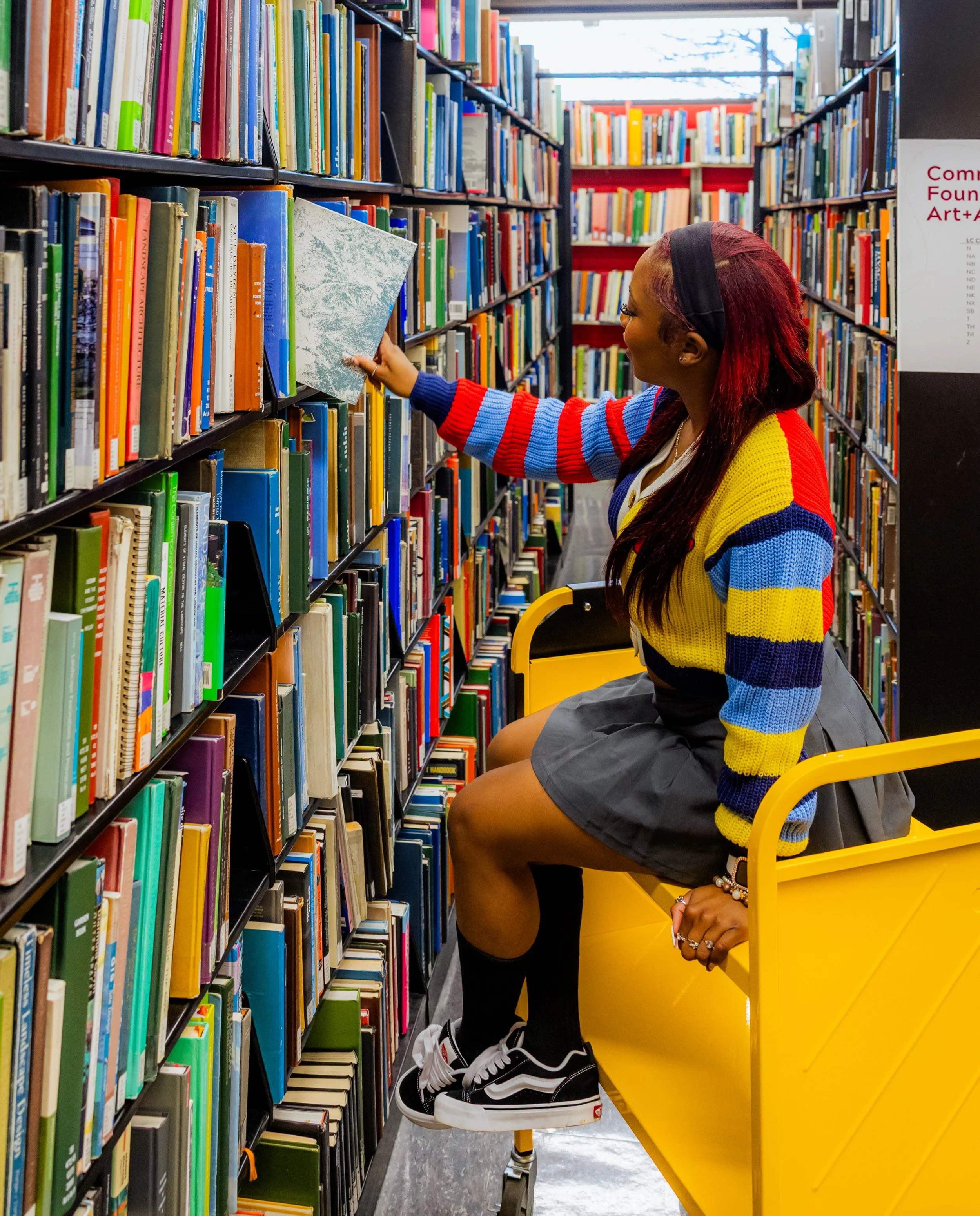 A woman browsing books in a library, sitting on a yellow cart, reaching for a book on a shelf.