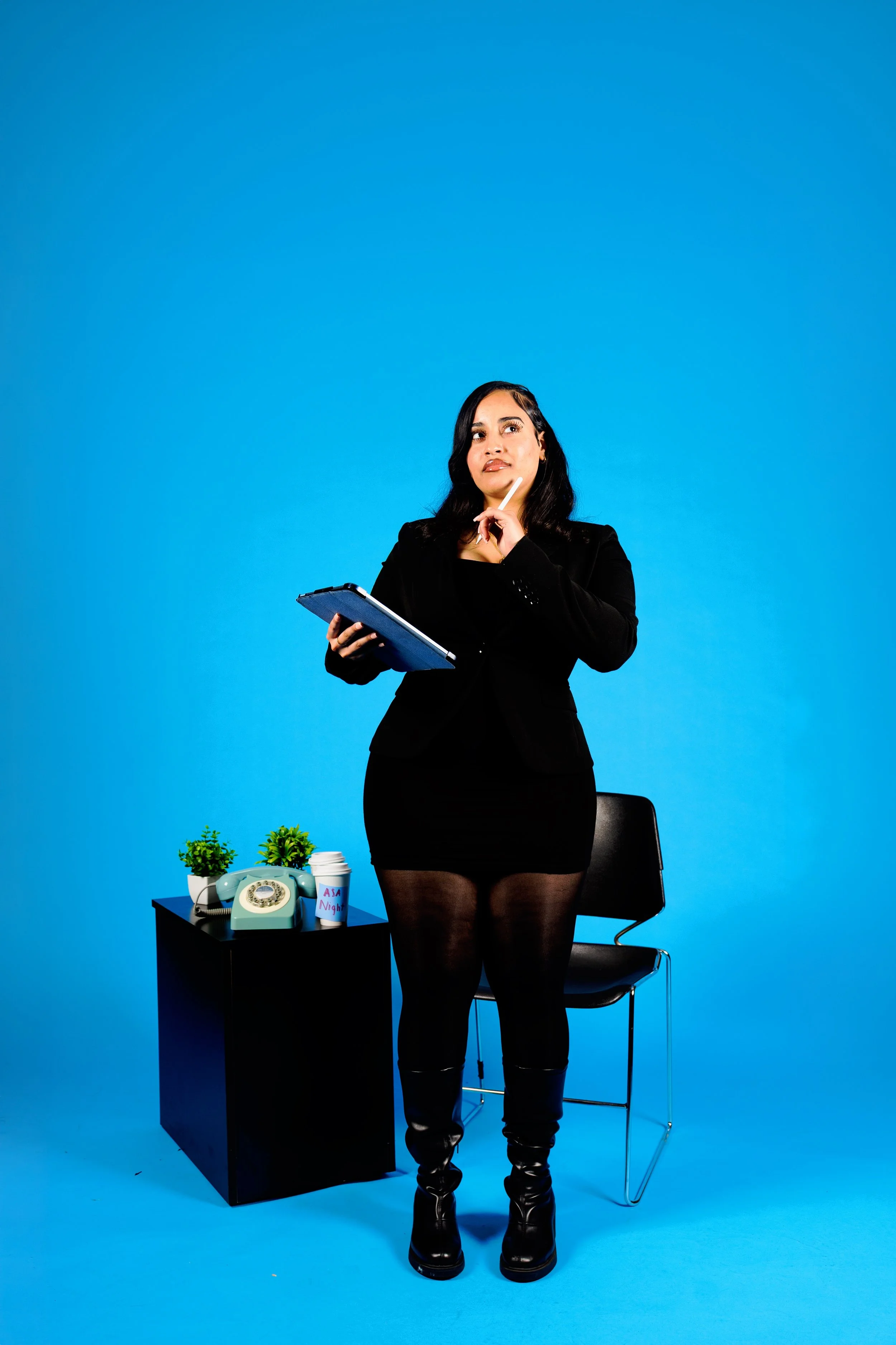 A woman dressed in black with a clipboard, standing against a bright blue background, with a black desk and chair nearby. The desk has two small potted plants, a vintage rotary phone, a cup labeled 'A Night', and a landline phone.