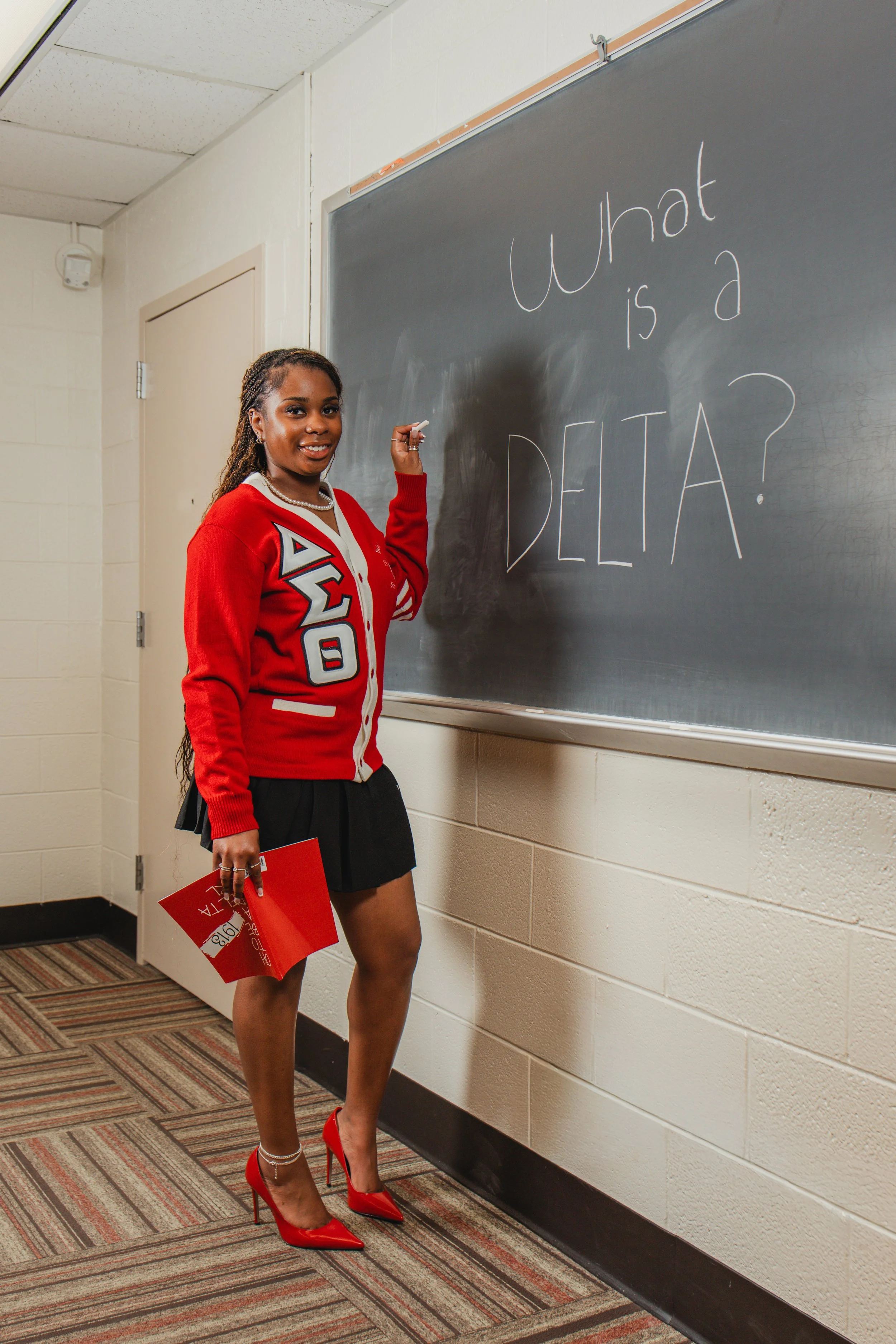 A young woman in a red sorority jacket, black skirt, and red high heels standing in a hallway next to a black chalkboard, smiling at the camera, holding a red folder, with the chalkboard reading 'What is a DELTA?'