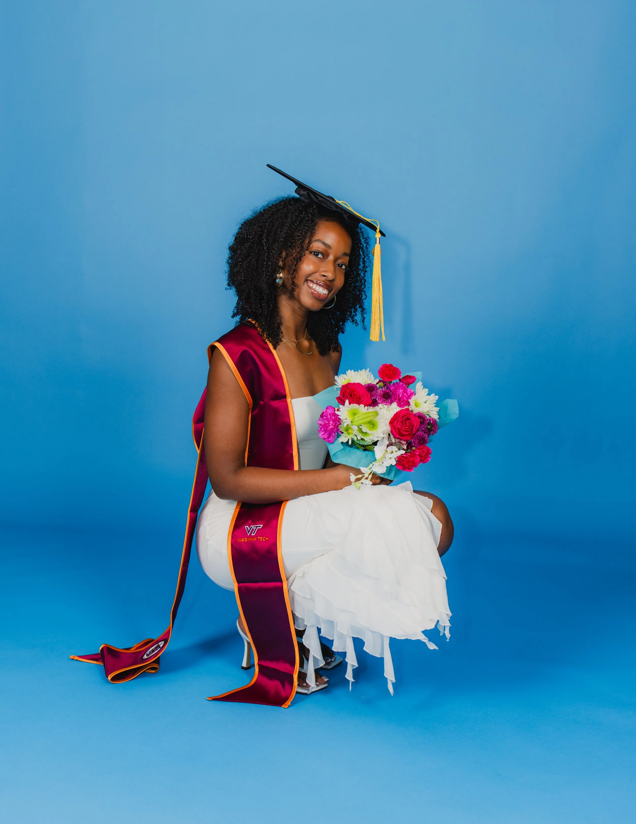 A young woman smiling in a graduation cap and gown holding a bouquet of flowers against a blue background.
