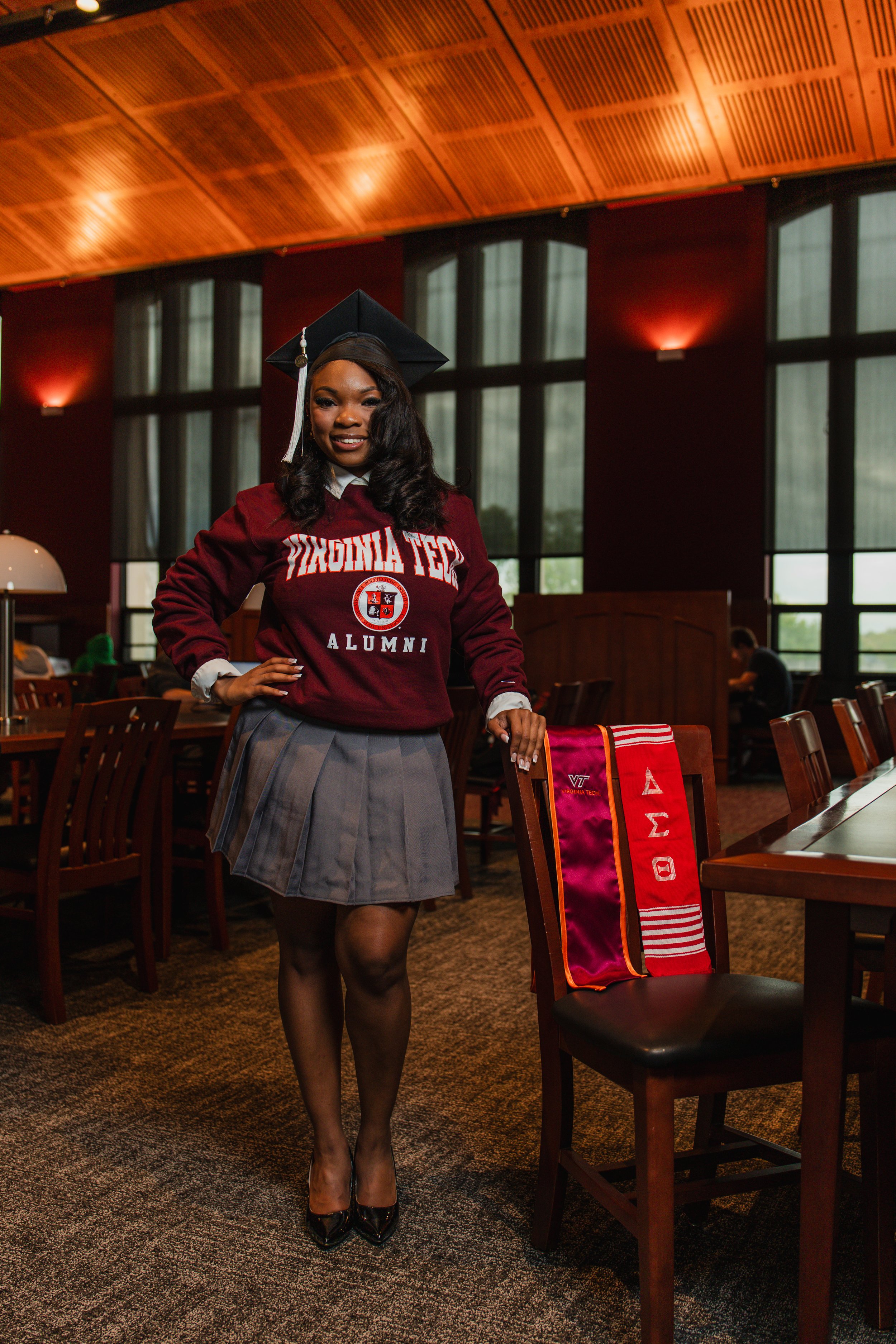 A woman in a graduation cap and gown with a 'Virginia Tech Alumni' sweatshirt, standing in a room with large windows and wooden ceiling, smiling at the camera.