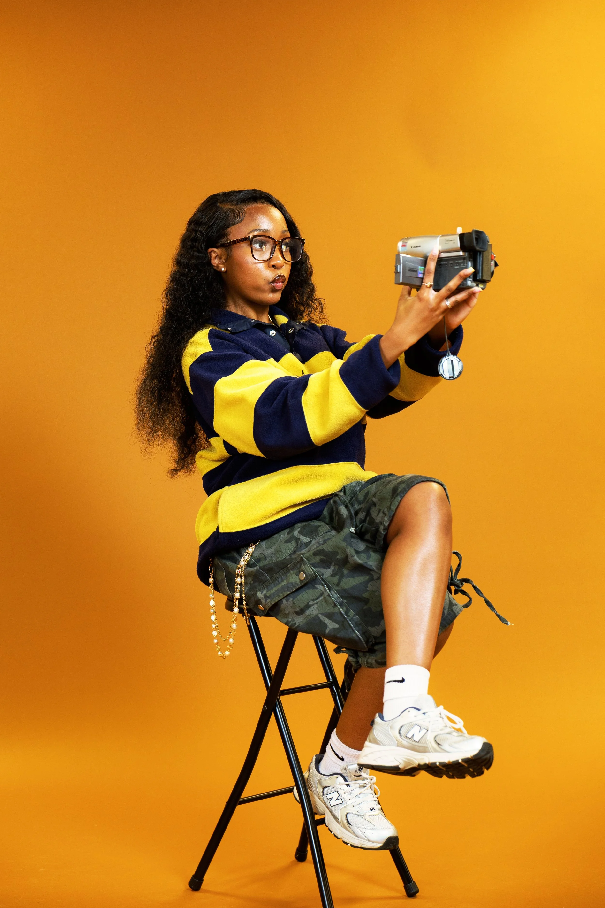 A young woman with glasses sitting on a black folding chair against a solid orange background. She is wearing a navy and yellow striped pullover, camouflage shorts, white socks, and white athletic shoes, and she is holding a vintage video camera.