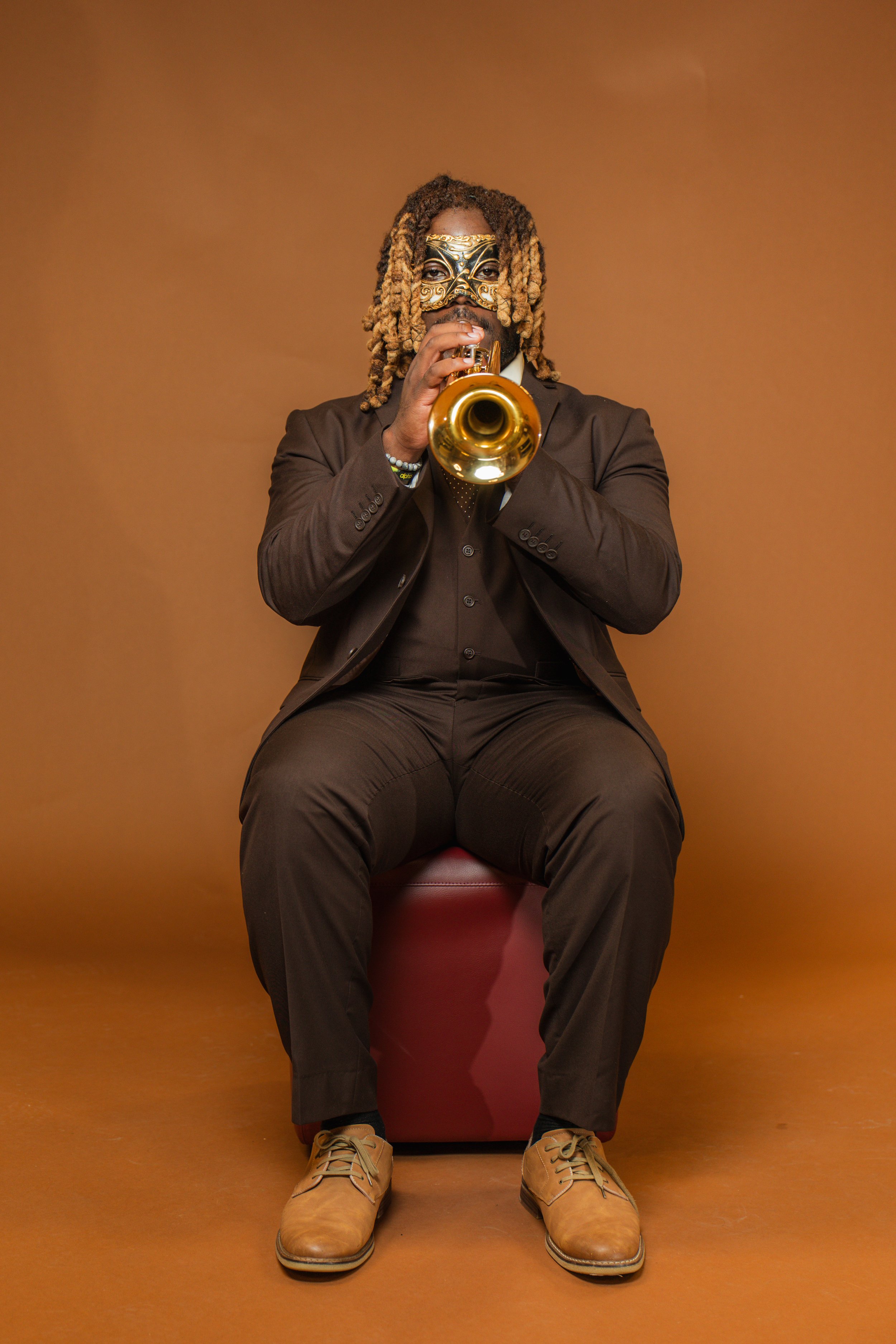 A person with long, blonde curly hair wearing a gold and black mask, a dark suit, tan shoes, and sitting on a red stool against a plain brown background, is playing a trumpet.
