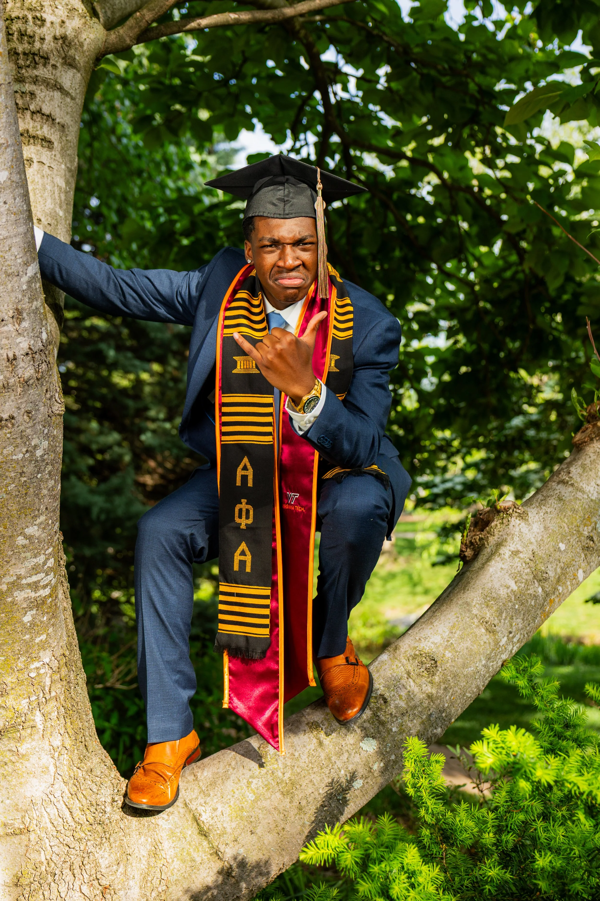 A young man wearing a graduation cap and gown with honors stole, sitting on a tree branch, making a silly face, dressed in formal attire with brown shoes, in a lush green outdoor setting.