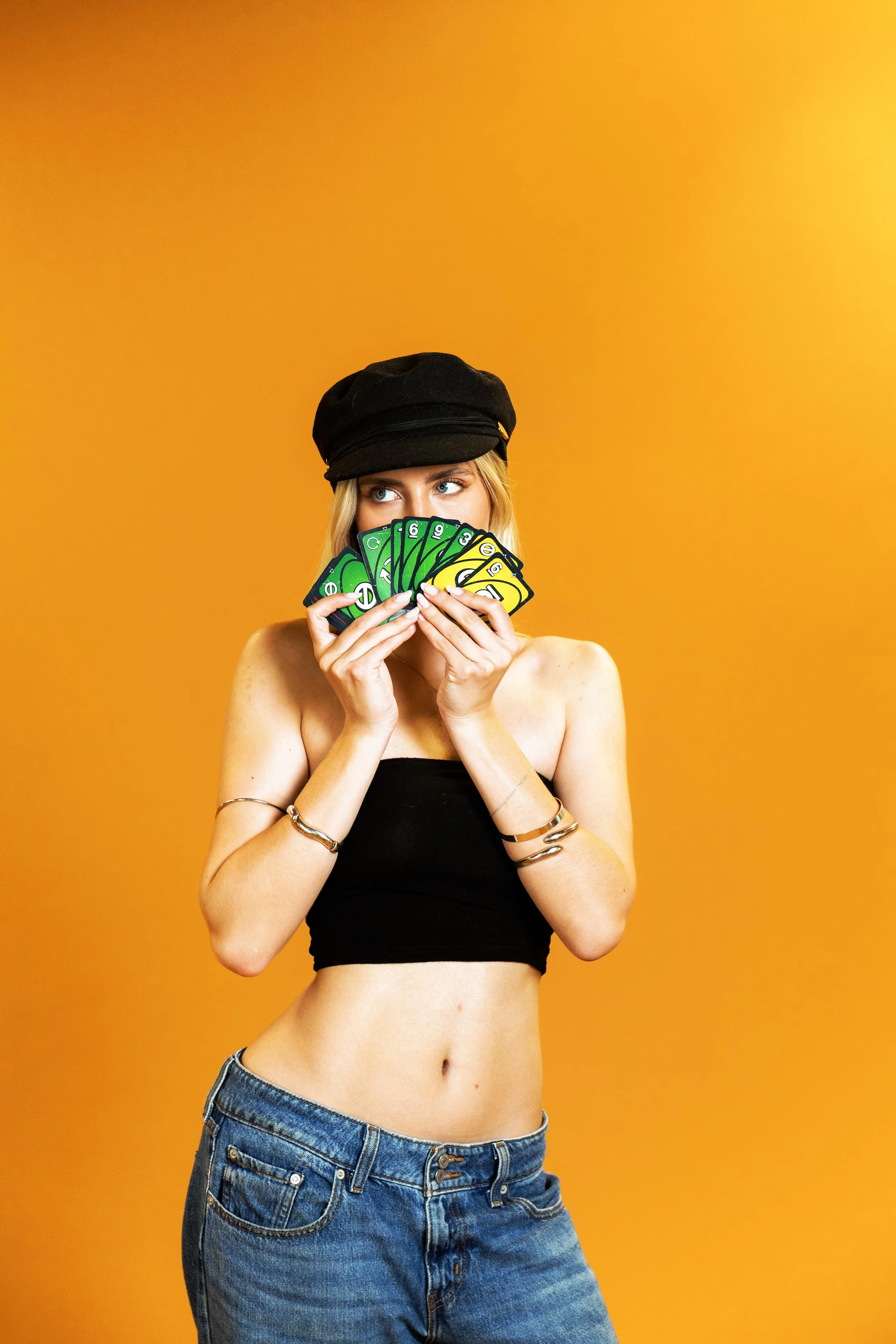 Young woman wearing a black cap and black tube top, holding a fan of UNO cards in front of her face, standing against an orange background.
