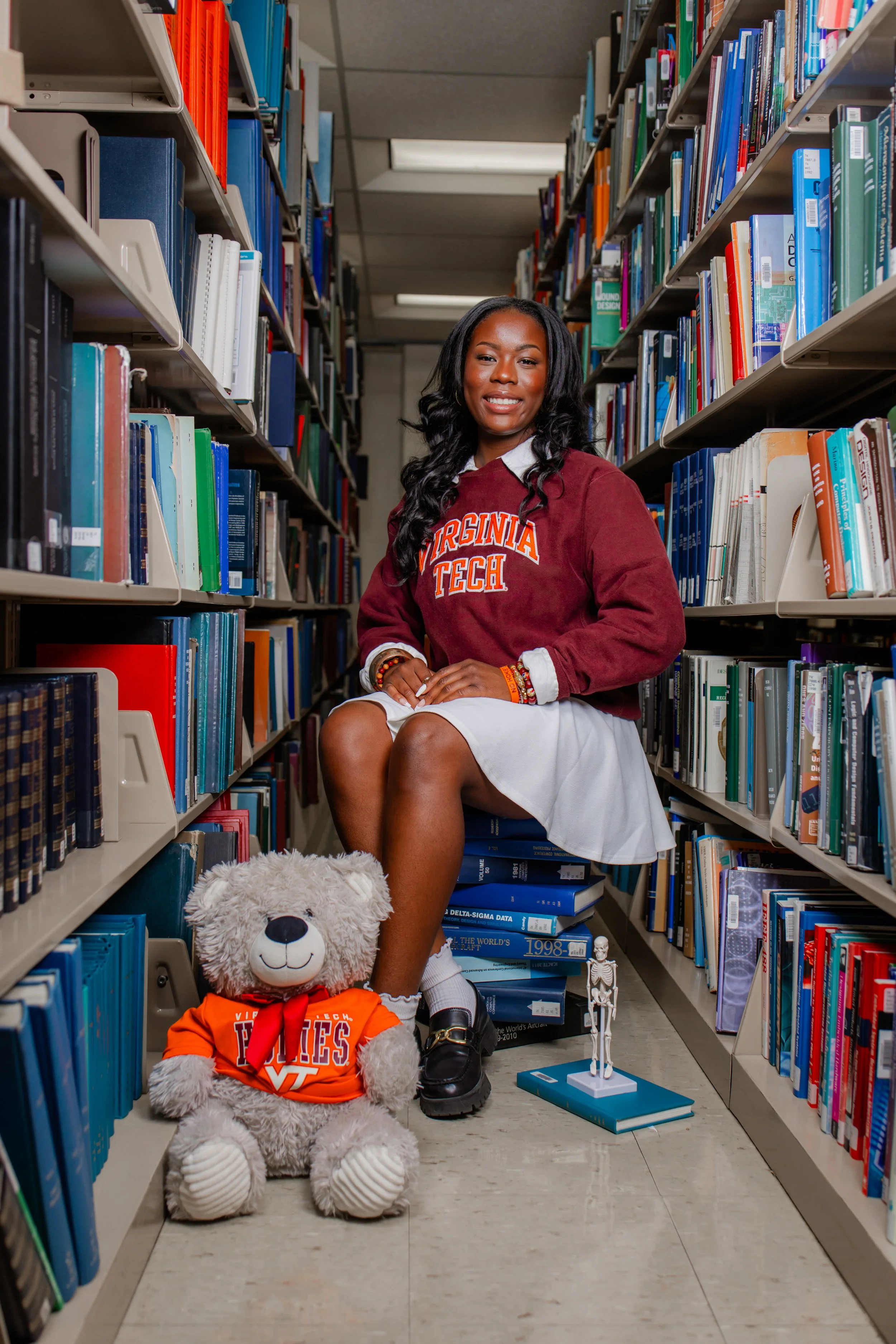 A woman sitting in a library aisle with a big teddy bear at her feet, wearing Virginia Tech apparel. The aisle has shelves filled with books, and there are small decorative skeleton and globe models on the floor nearby.