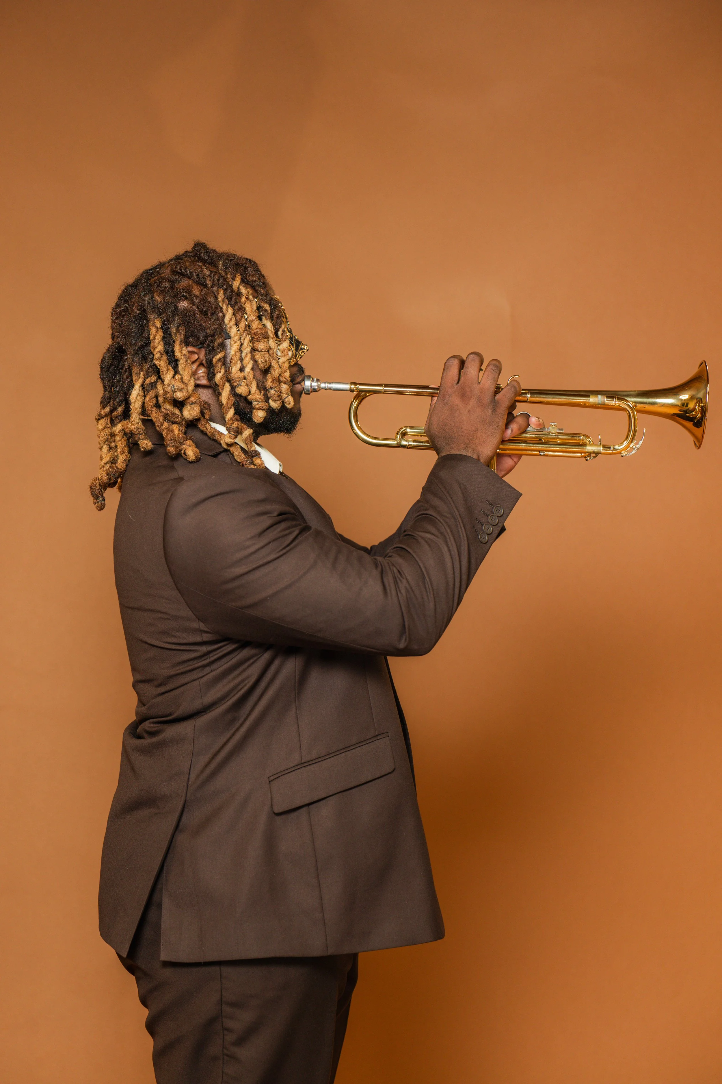 A man with dreadlocks in a brown suit playing a trumpet against a brown background.