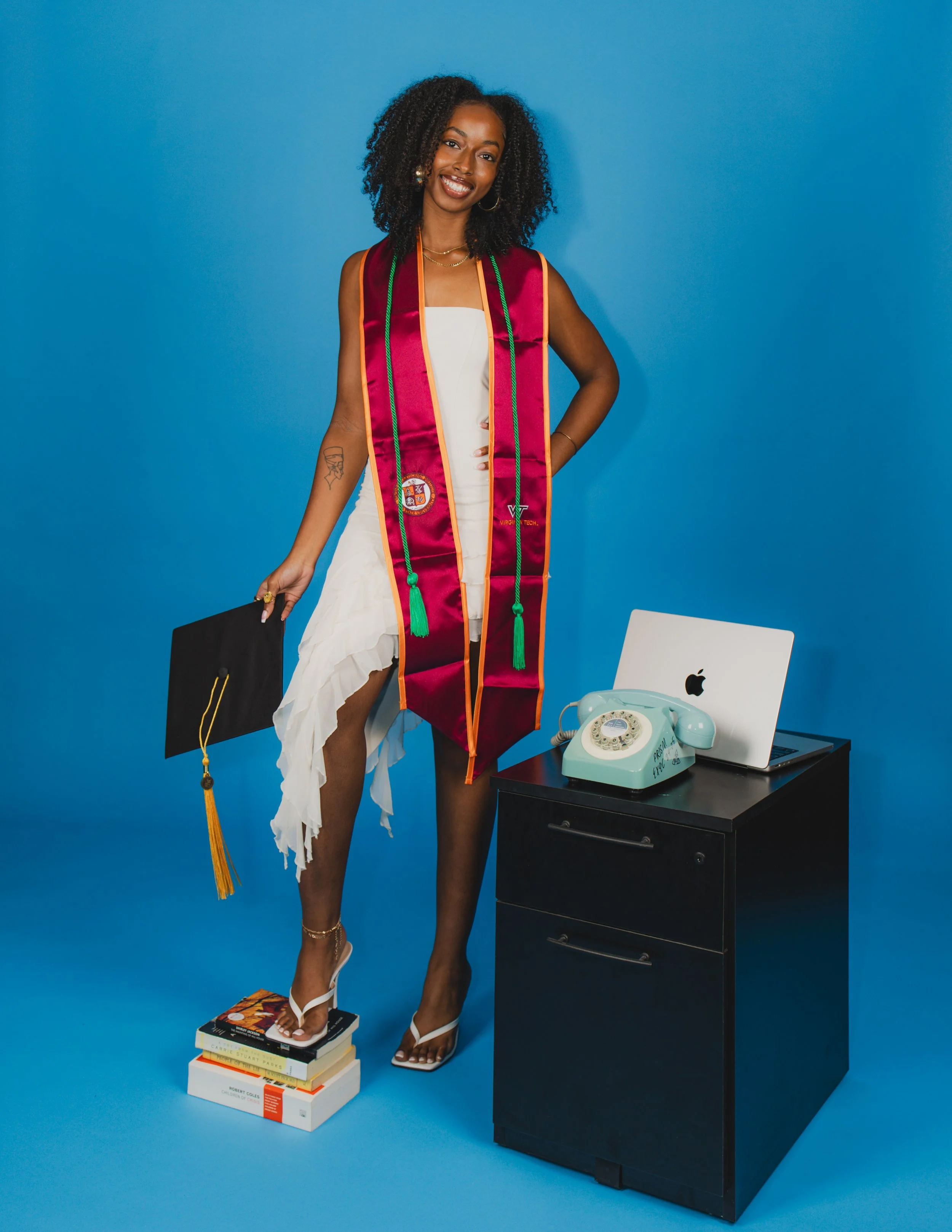A woman in a white dress wearing a graduation stole and standing on a stack of books, with her right hand on her hip, smiling against a blue background with a black desk, a vintage rotary phone, a MacBook, and a turquoise mug on it.