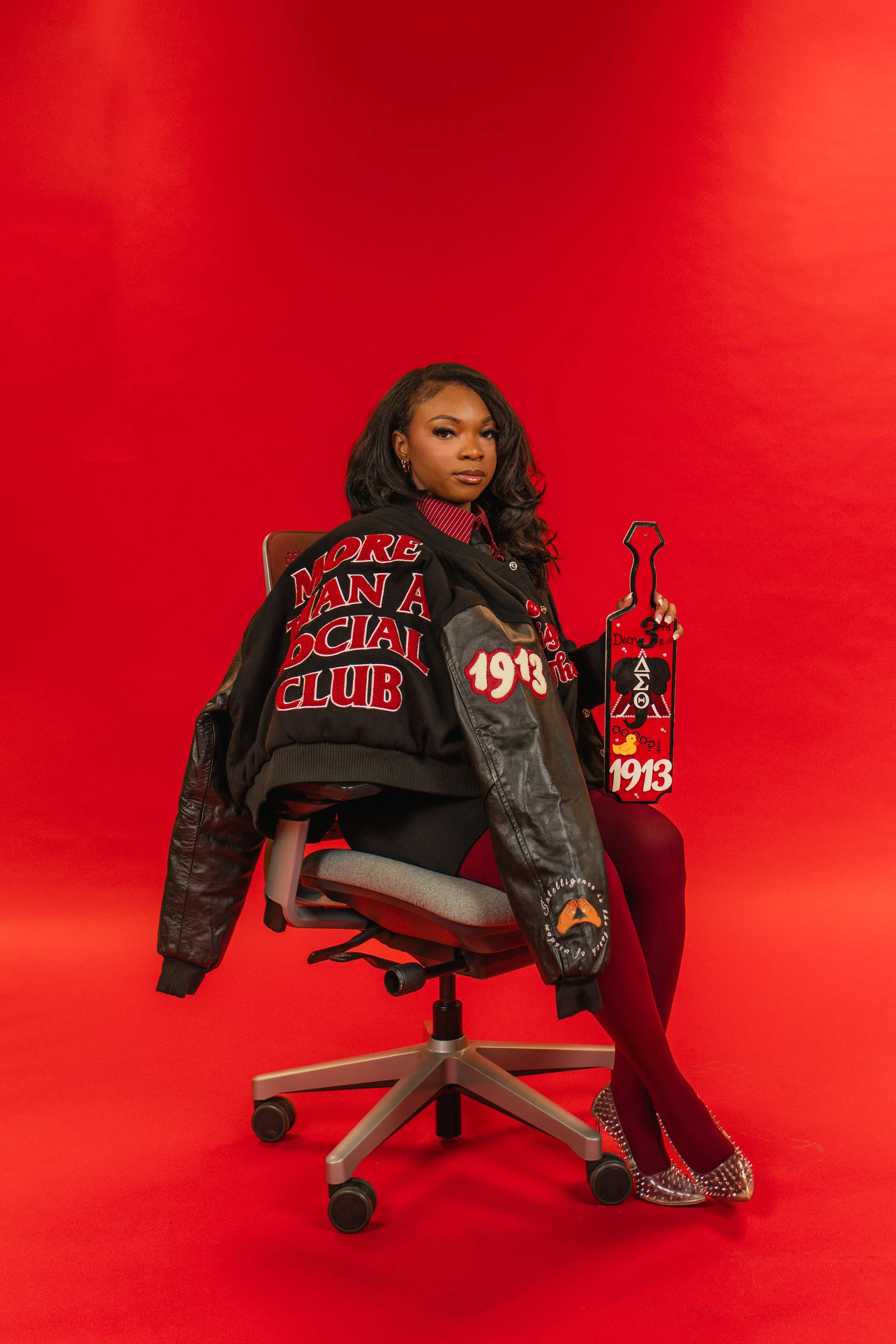 A woman sitting on an office chair against a red background, wearing a black jacket with patches and lettering, colorful high heels, and holding a decorated red and black scroll. The chair's wheels are visible, and she is looking confidently at the c