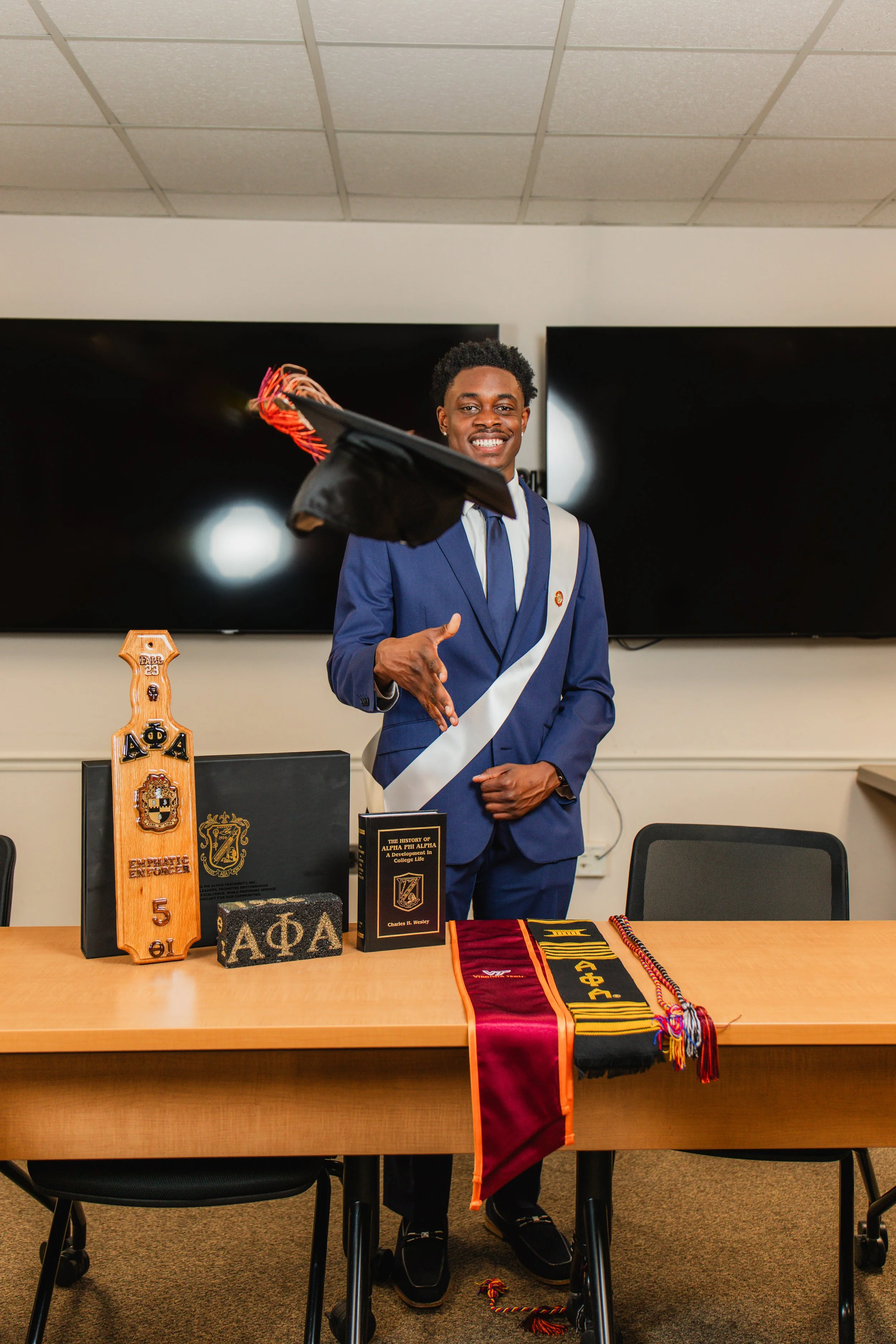 A young man in a blue suit and graduation cap is standing behind a table, smiling and pointing. The table displays various fraternity memorabilia, including banners, cords, and trophies. Large black screens are mounted on the wall behind him.