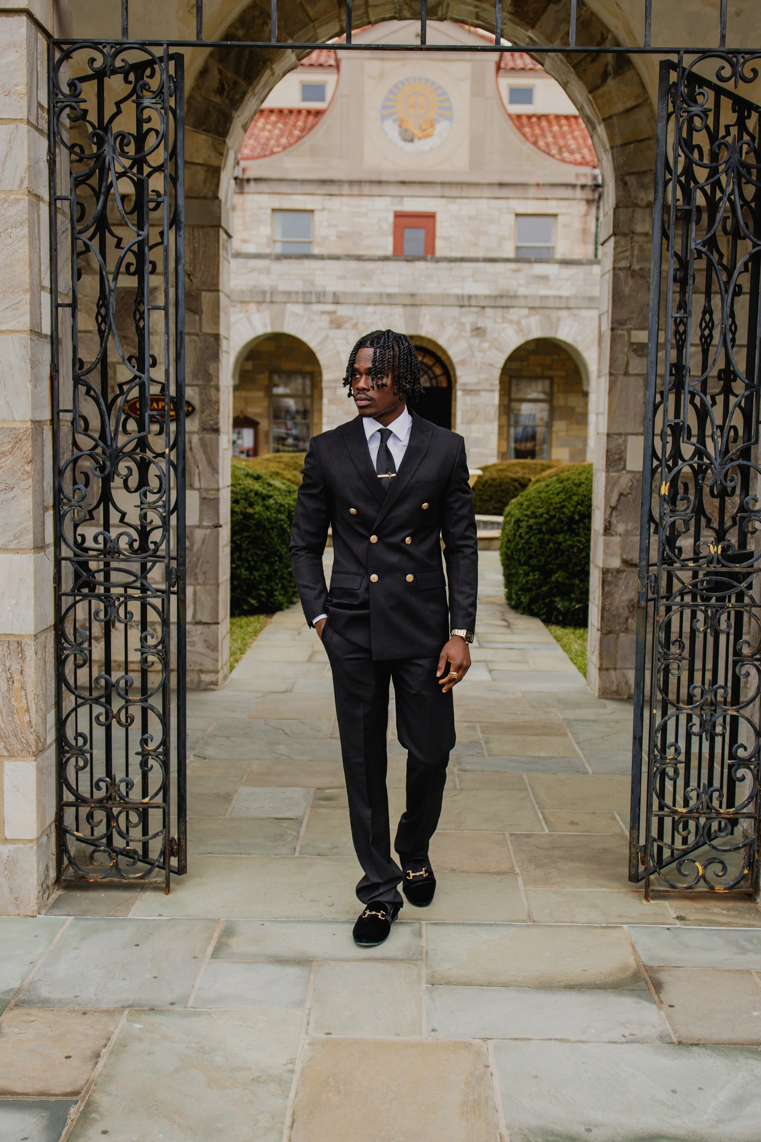 A young man in a black suit, white shirt, and black tie walking through an ornate iron gate with a stone building and landscaped bushes in the background.