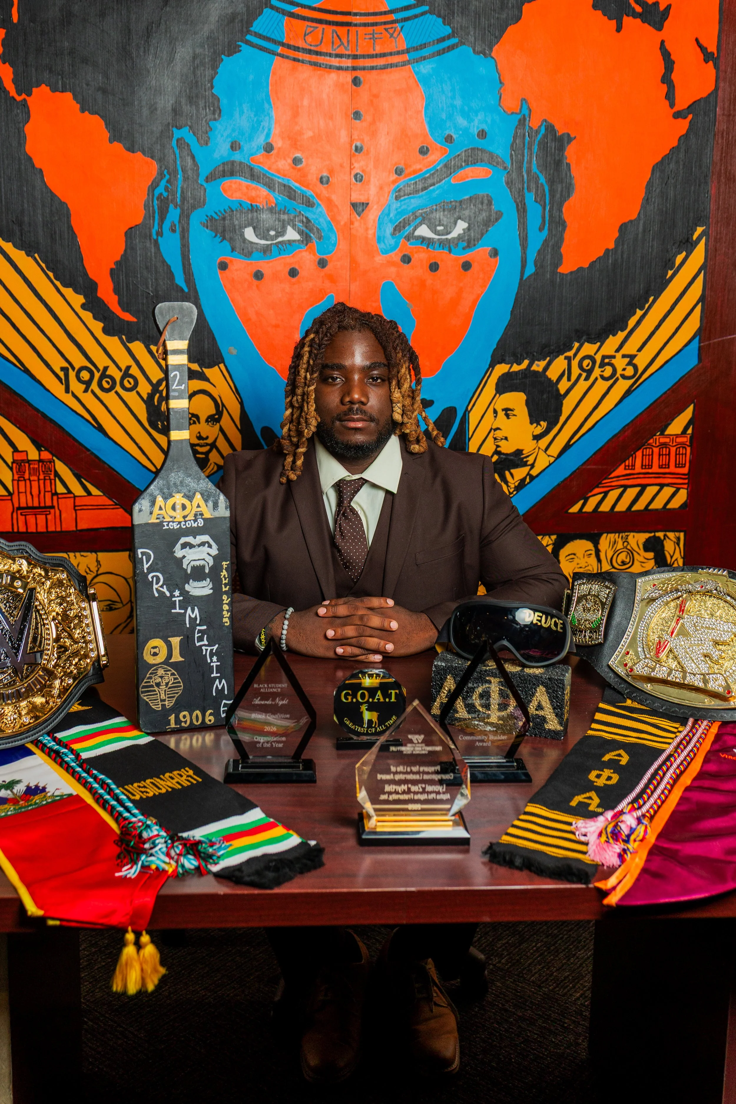 A man with long twisted hair in a suit sitting at a table with awards, trophies, and colorful cloths, in front of a mural of a face with orange and blue colors and portraits of people from the African American community.