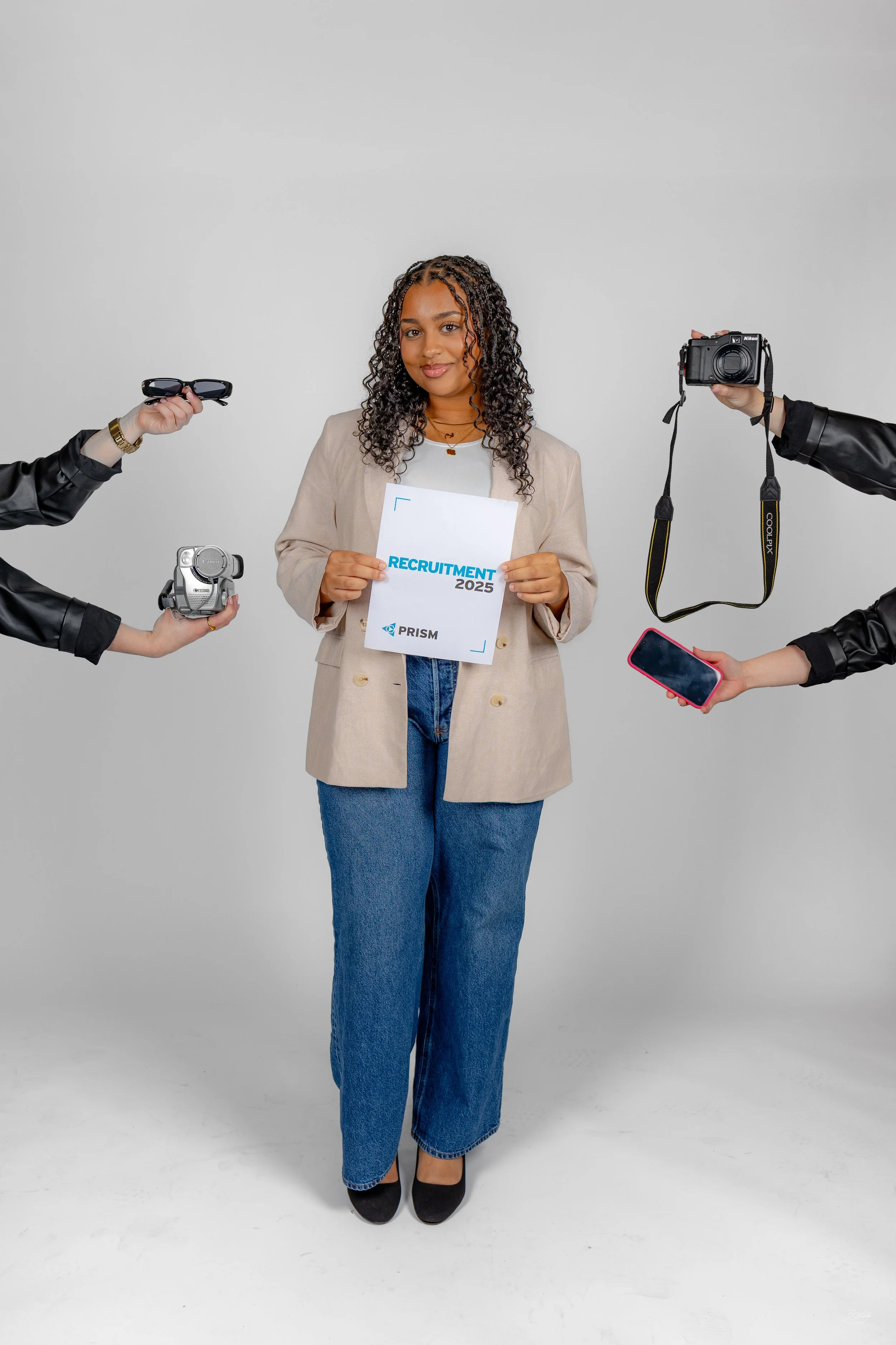 A woman with curly hair wearing a beige blazer, white shirt, and blue jeans, holding a document that says 'RECRUITMENT 2025' with the PRISM logo. She is surrounded by four hands holding various objects: sunglasses, a camera, a Zoom recorder, and a sm