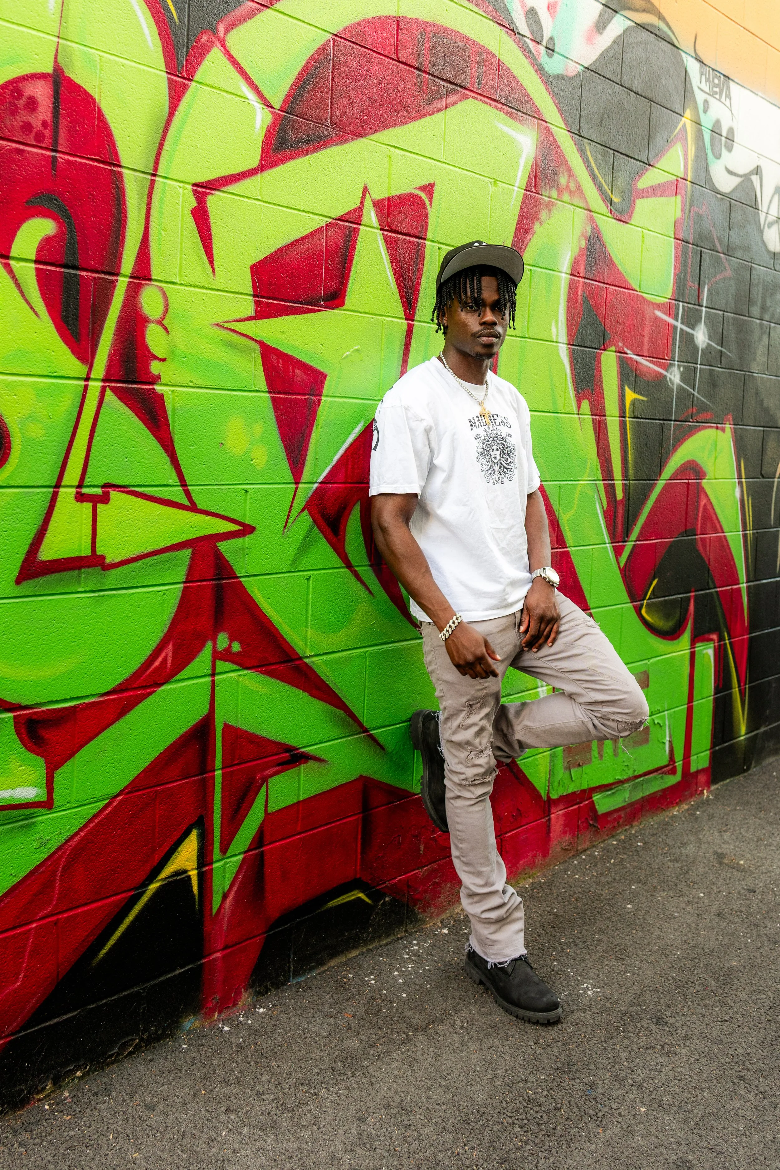 A young man with dreadlocks wearing a gray cap, white t-shirt, beige pants, and black shoes standing against a colorful graffiti wall with red, green, black, and yellow designs.
