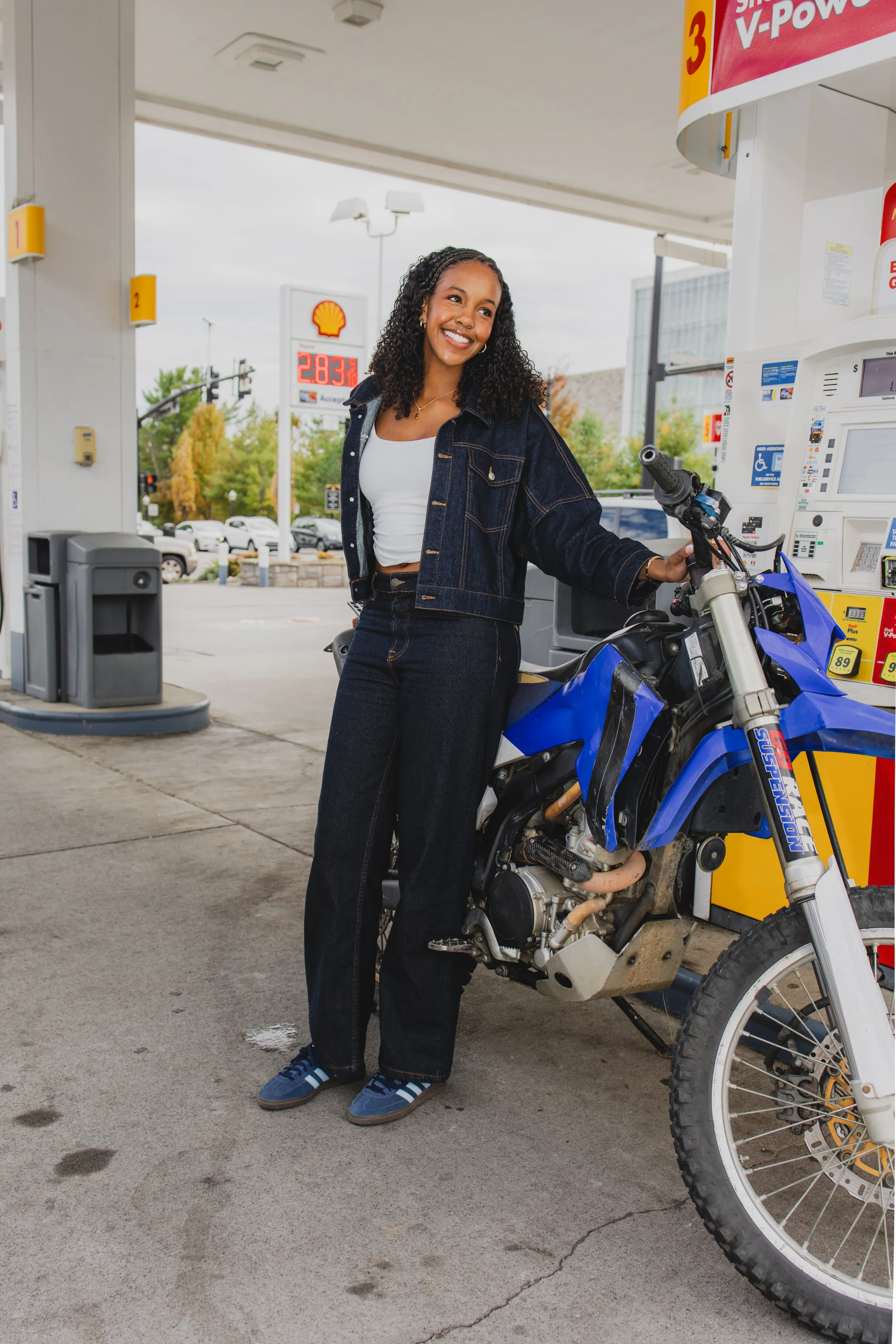 A smiling young woman with curly hair, wearing a denim jacket and jeans, standing next to a blue dirt bike at a Shell gas station. She is holding the handlebar of the motorcycle with her right hand. In the background, there are gas prices displayed o