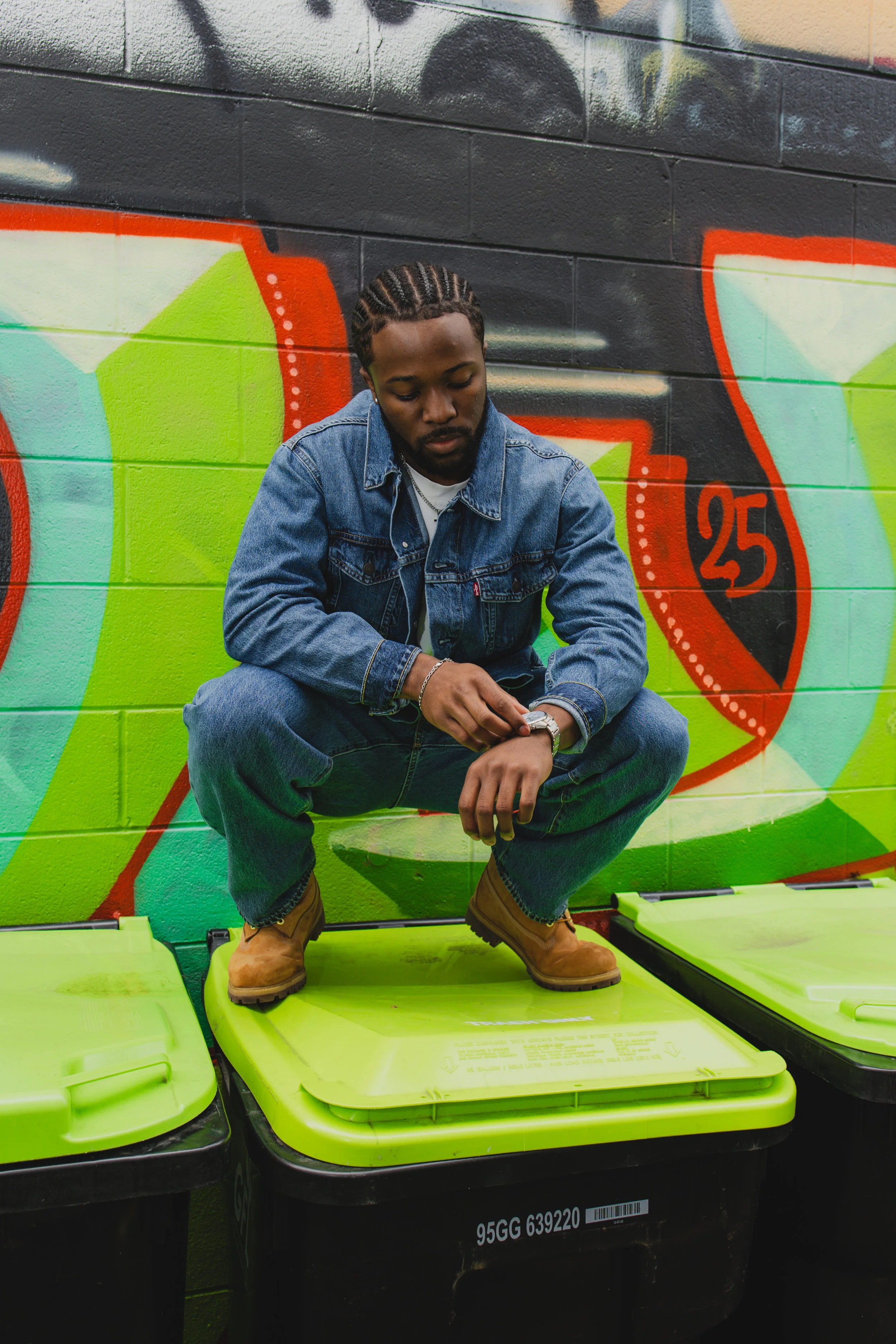 A young man with braided hair, wearing a denim jacket, sitting on a lime green trash bin in front of a colorful graffiti wall, looking at his wristwatch.