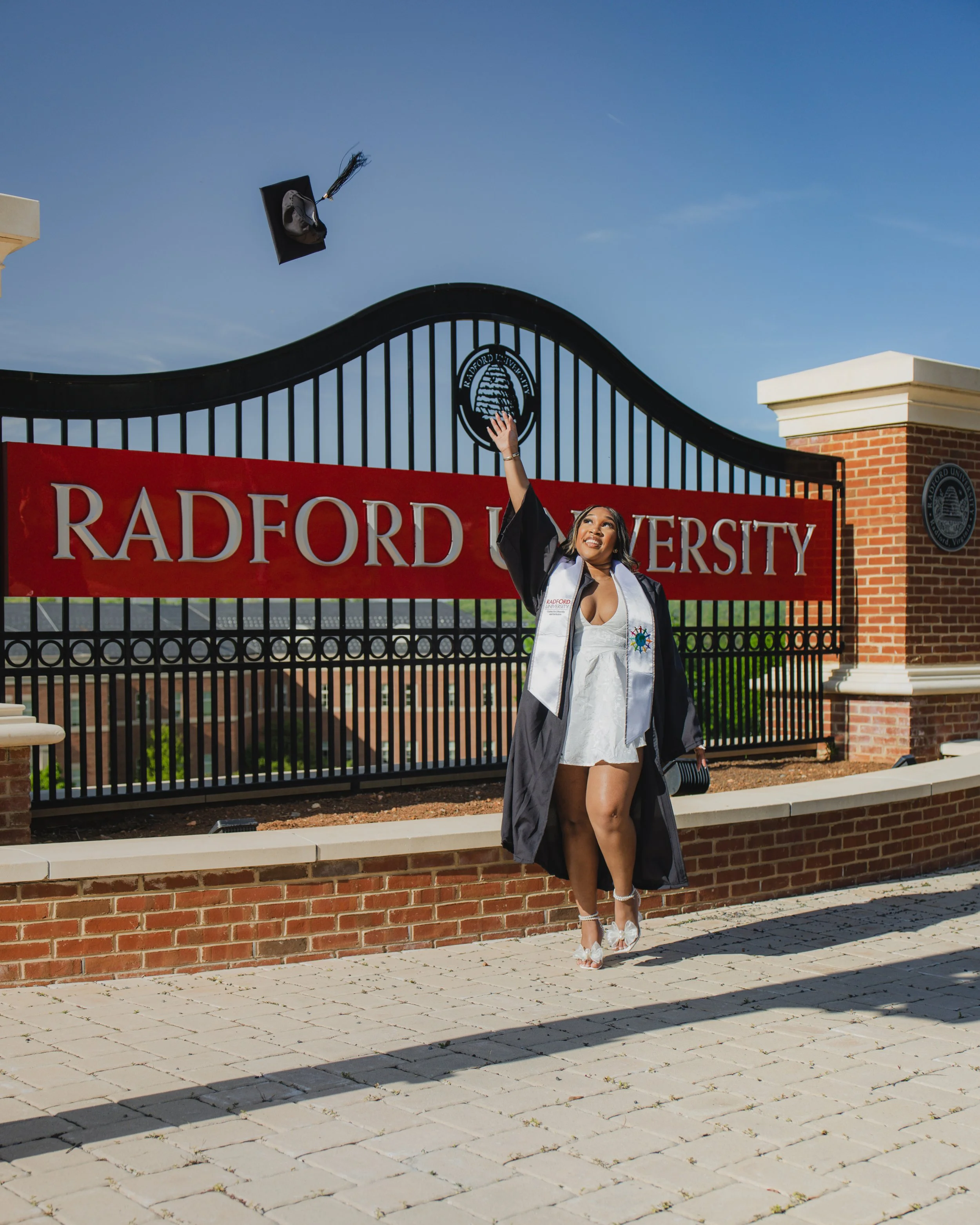 A young woman in a graduation gown and cap stands in front of a gate with a sign that reads "RAYFORD UNIVERSITY". She is smiling and throwing her cap into the air, celebrating her graduation on a sunny day with a clear blue sky.