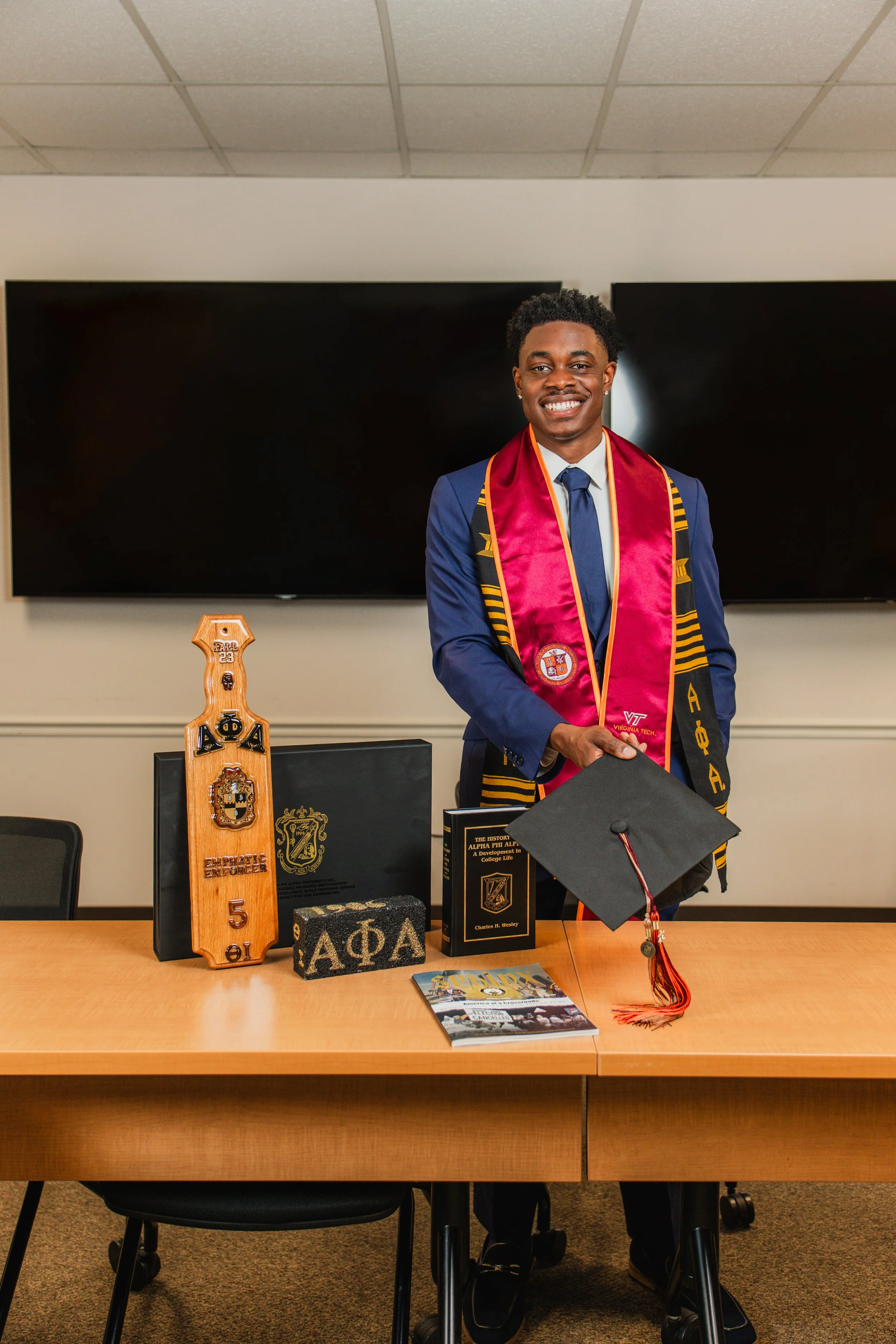 Young man in academic regalia smiling at graduation ceremony, standing behind a table with honorary items and publications, in a room with large digital screens on the wall.