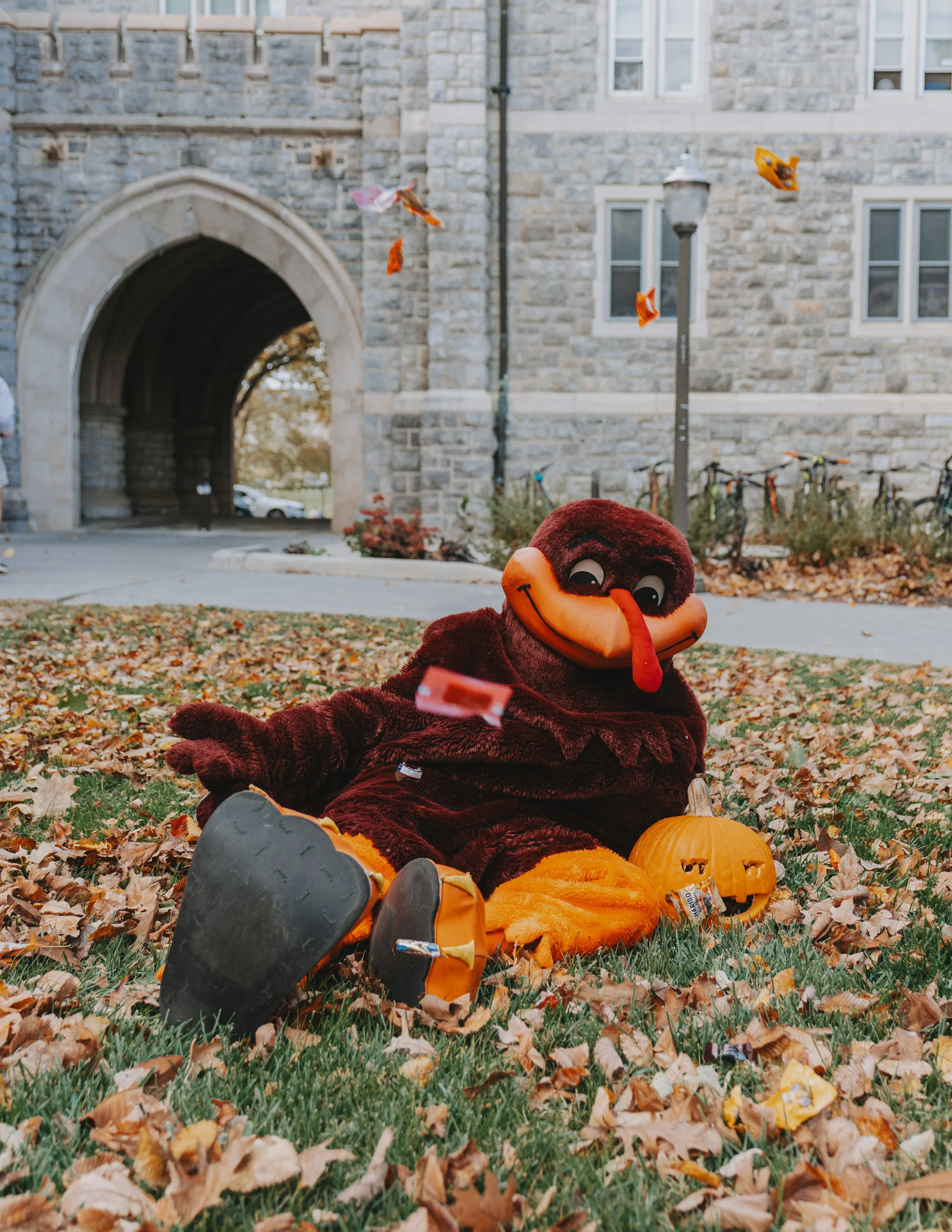 A person dressed in a turkey mascot costume sitting on the ground among fallen autumn leaves, with a carved pumpkin next to them, in front of a stone building and a lamppost, during Halloween.