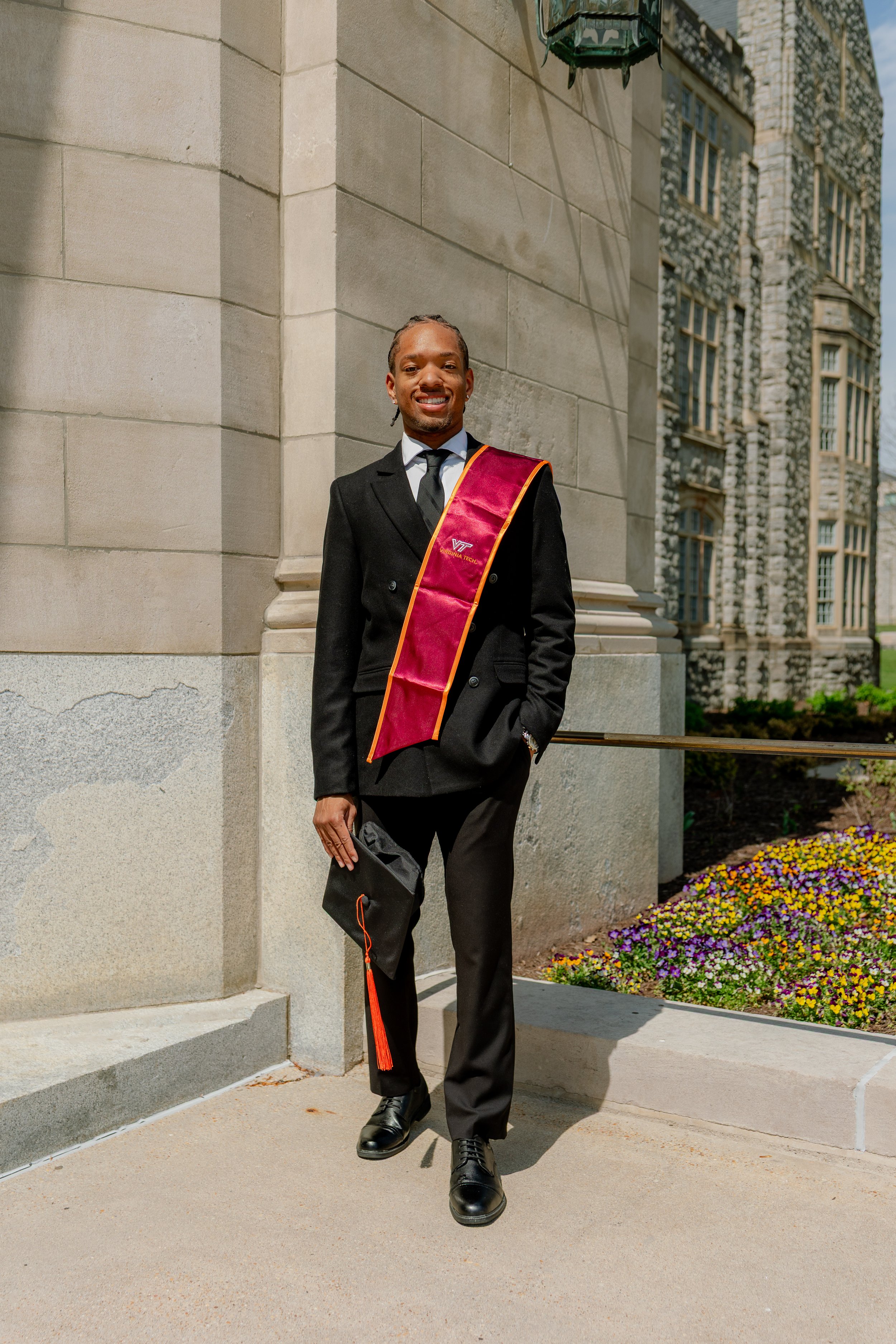 A young man dressed in a black suit with a Virginia Tech graduation stole holds a graduation cap and hangs out of frame, standing outdoors near a stone building and colorful flower beds.