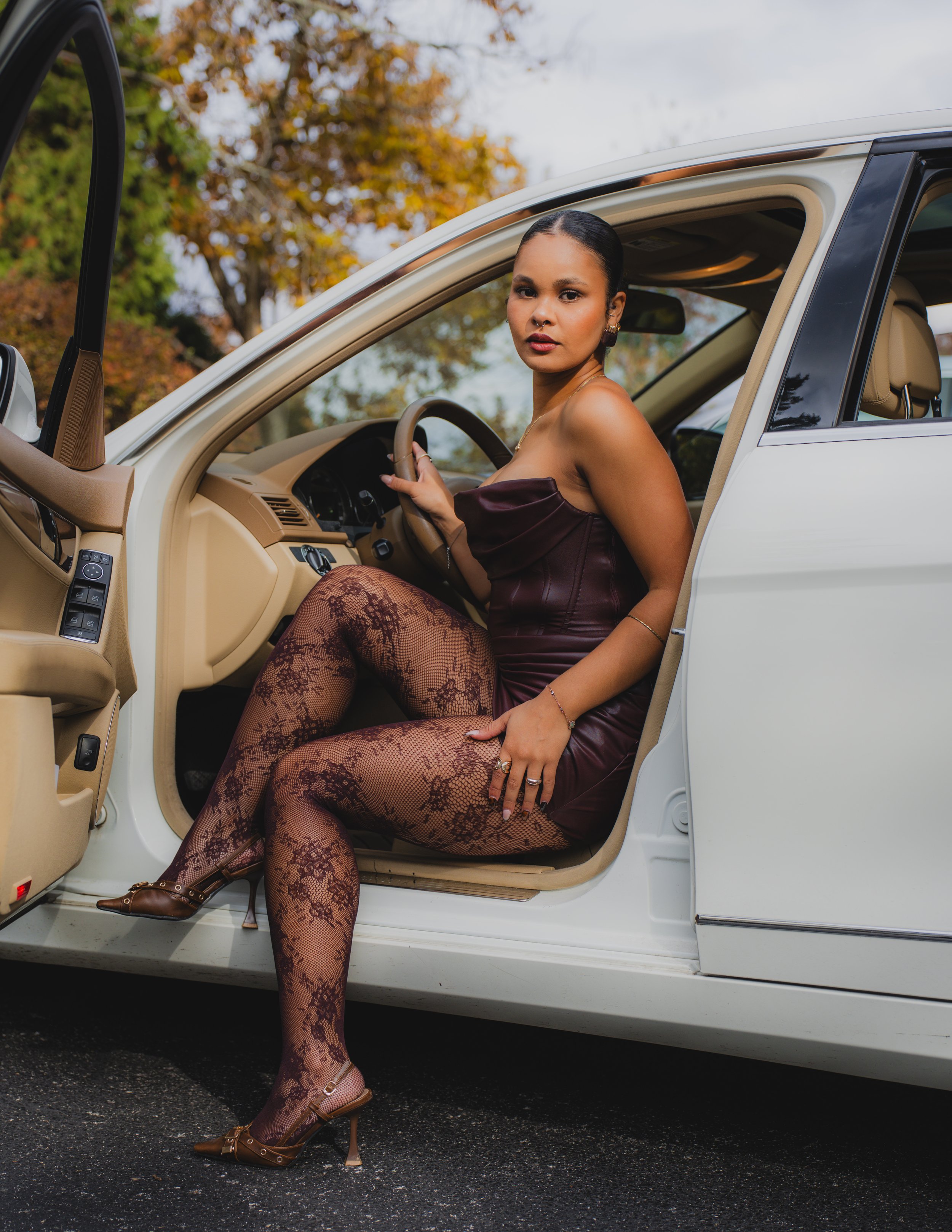 A woman sitting in the driver's seat of a white car, wearing a strapless dress, lace tights, and high heels, outdoors with trees in the background.