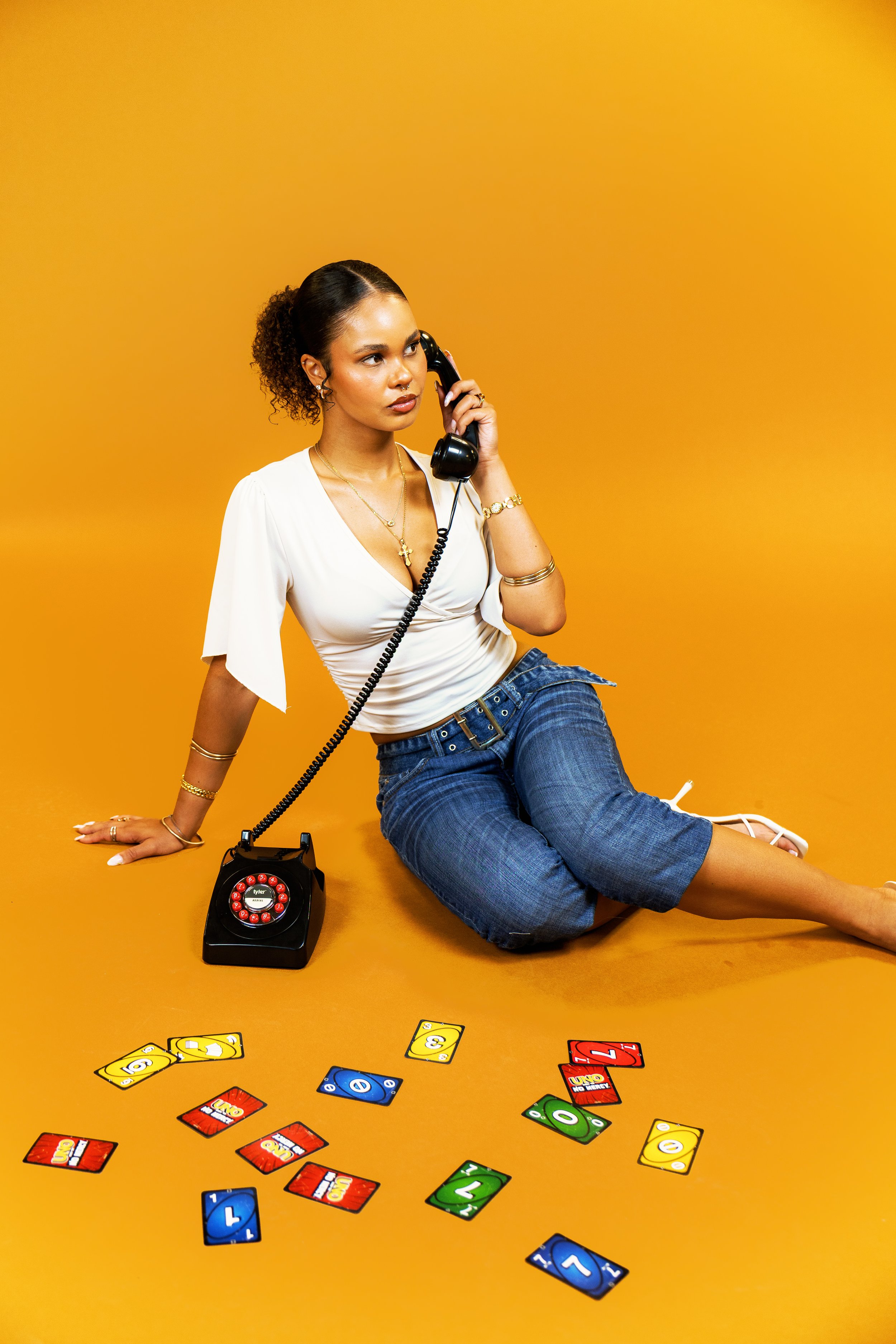 A woman with curly hair tied back, holding a vintage black rotary phone to her ear, sitting on a yellow background. There are Uno cards and a black rotary dial phone on the floor in front of her.