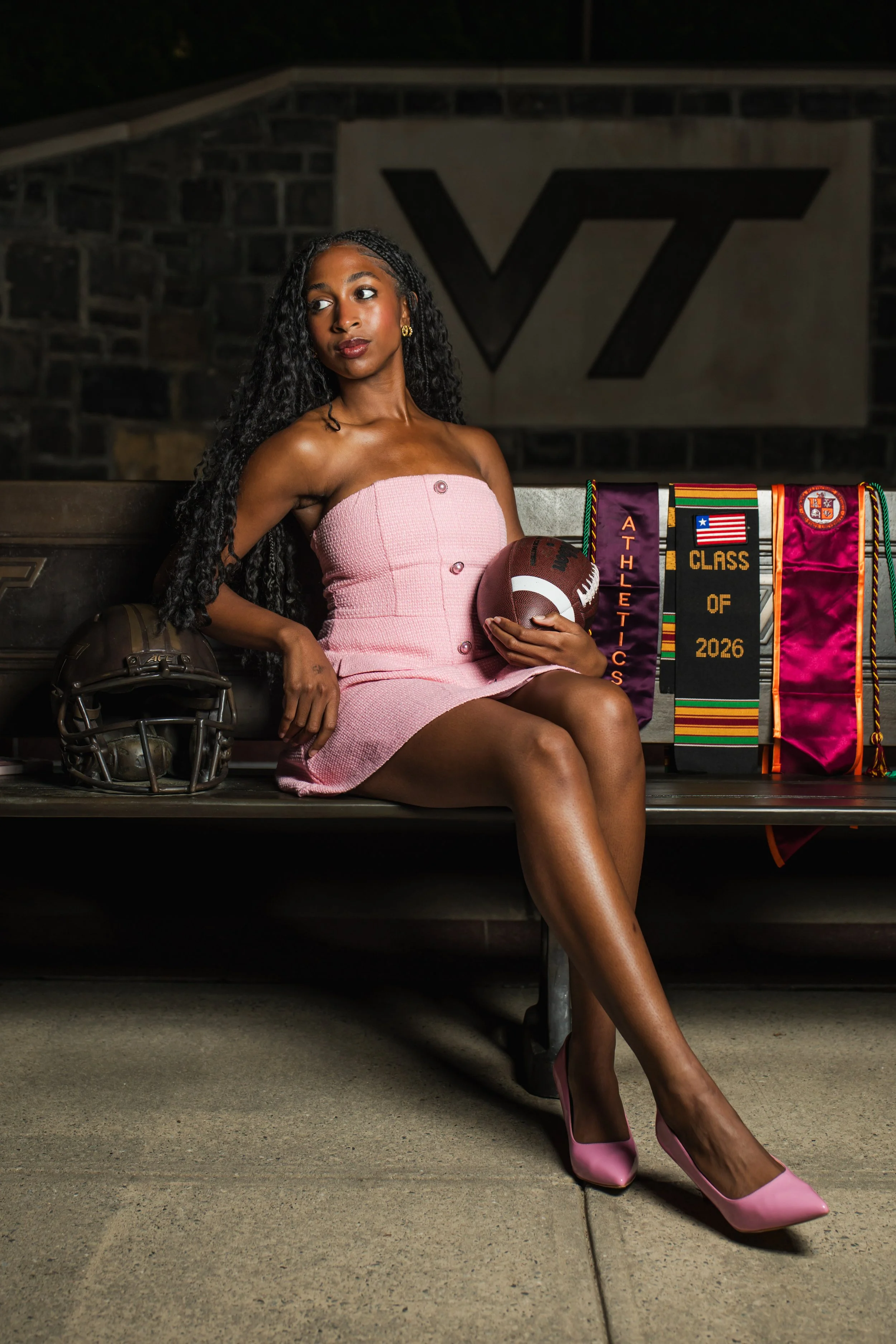 A woman with long curly black hair sitting on a bench at night. She is wearing a pink strapless dress, pink high heels, and gold earrings. She is holding a football in her left hand. There are colorful awards or ribbons hanging on the side of the ben