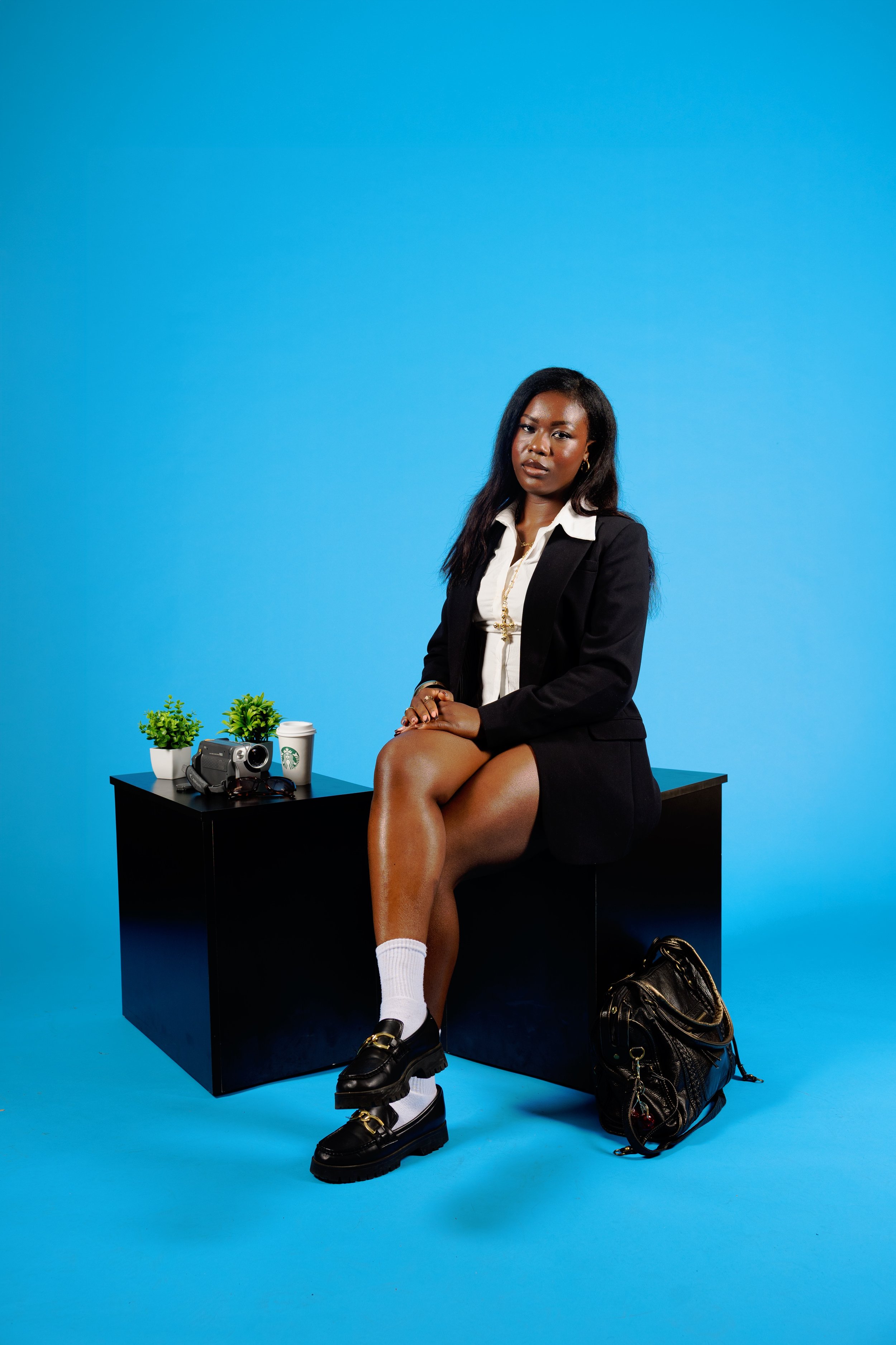 A woman sitting on a black table with her legs crossed against a blue background. She is wearing a black blazer, white shirt, black shoes, and white socks. There are two small green plants, a black video camera, and a Starbucks cup on the table. A bl