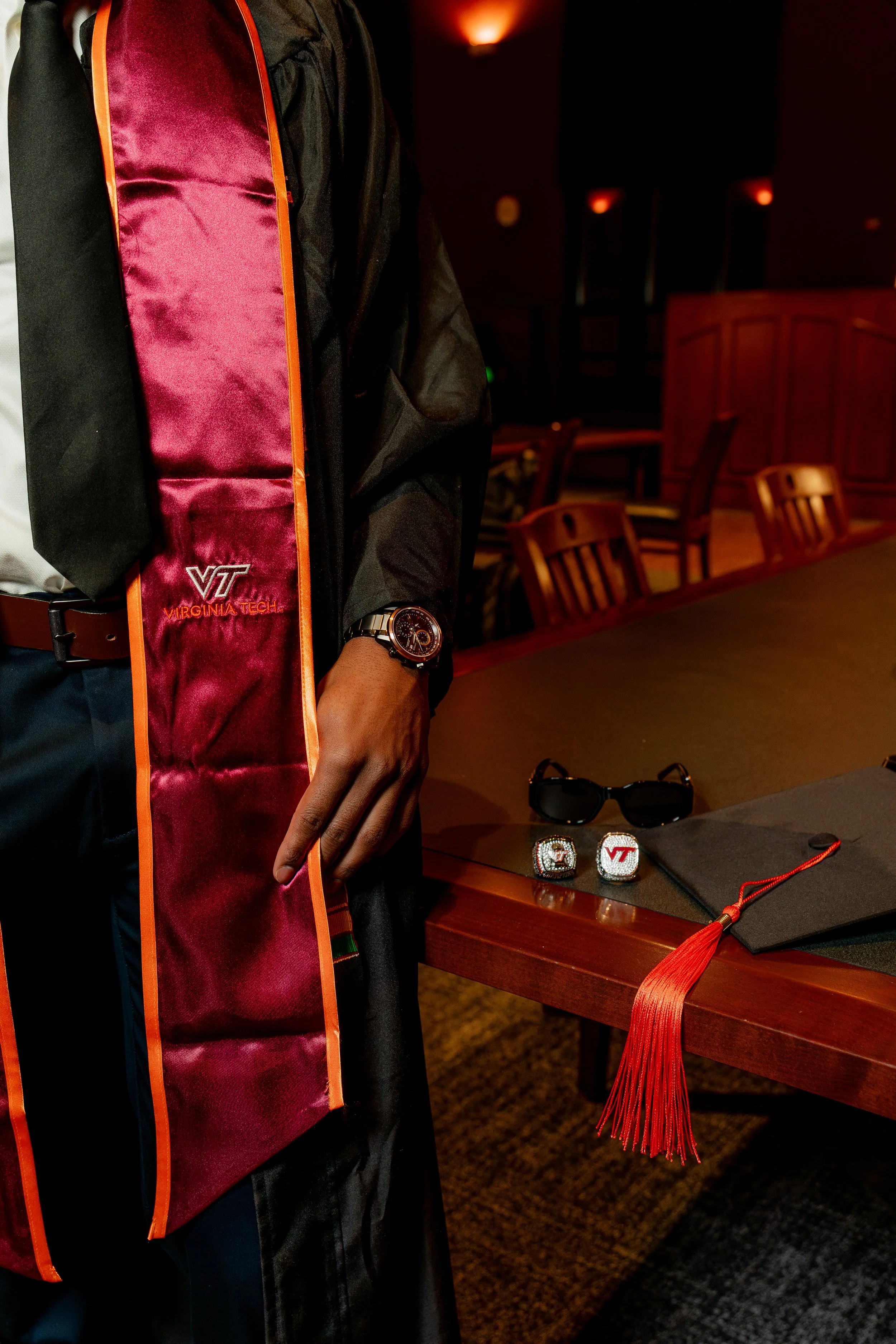 Man wearing a black gown with Virginia Tech graduation stole, watch, and rings, standing next to a table with graduation cap, sunglasses, and tassel in a dimly lit room.