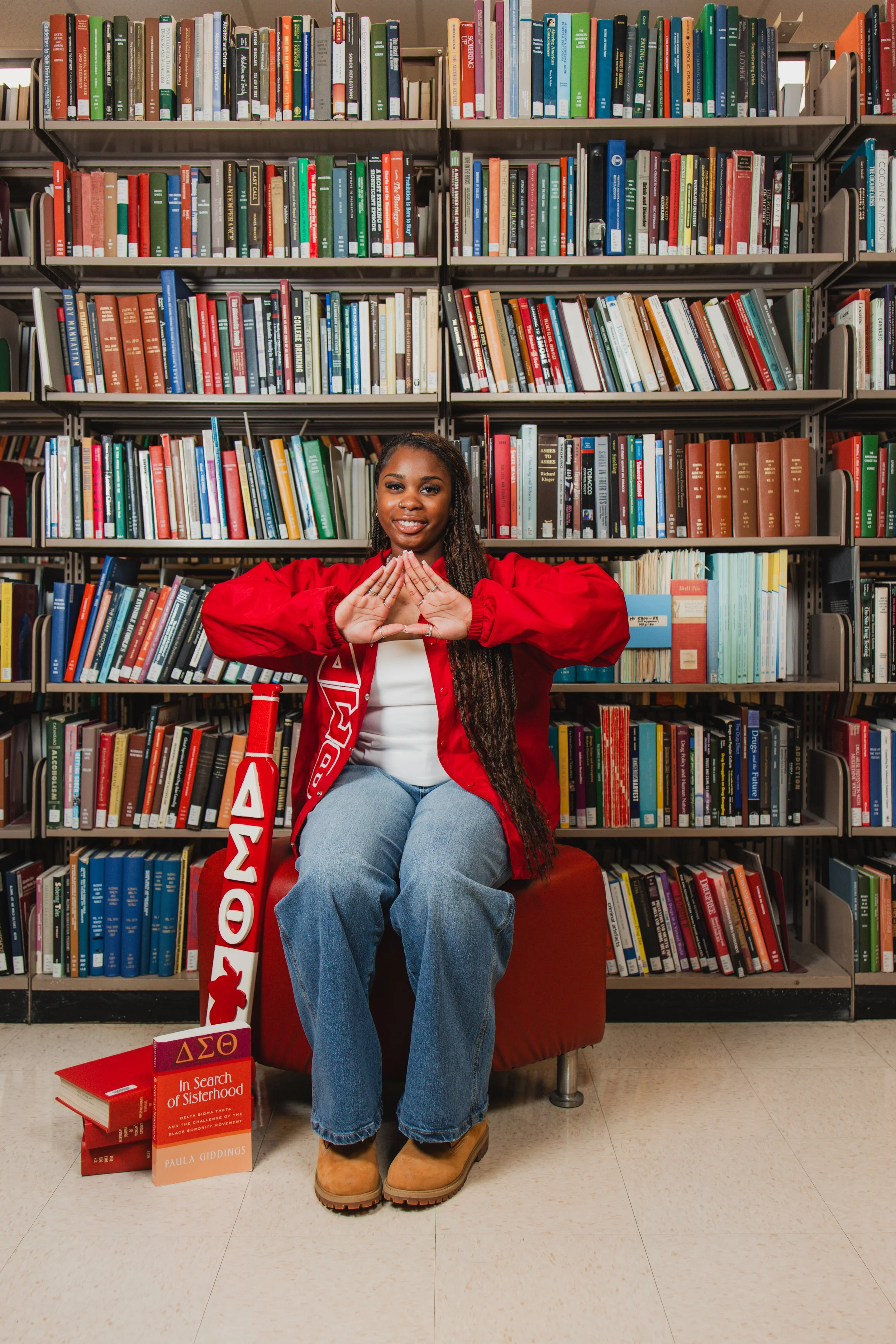 A young woman sitting on a red chair in a library, surrounded by books and holding her hands in a diamond shape in front of her chest. She is smiling and wearing a red jacket, blue jeans, and tan boots, with books about sisterhood and social movement