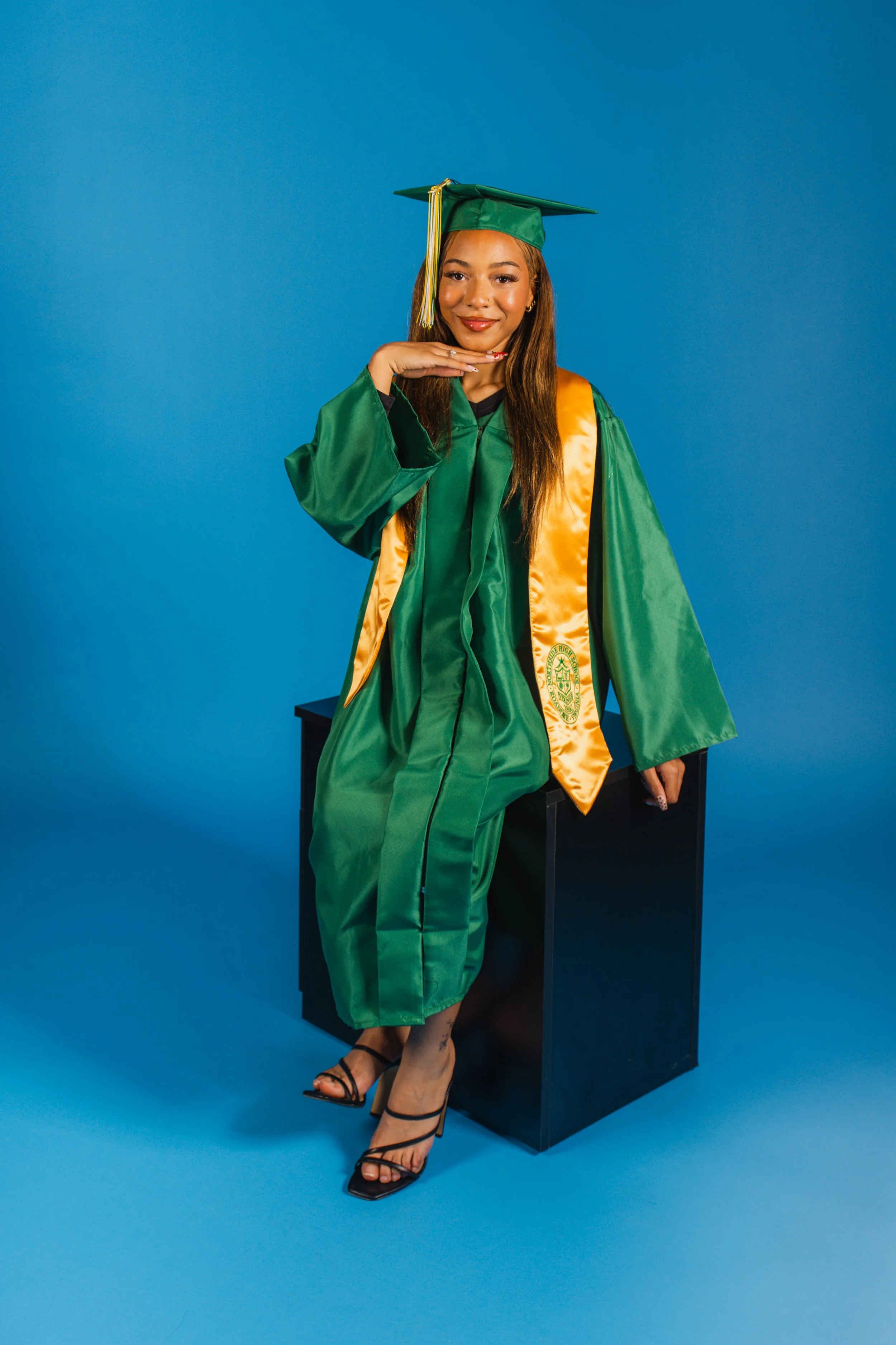 Young woman in a green graduation gown and cap with gold accents, sitting on a black pedestal against a blue background, smiling and posing confidently.