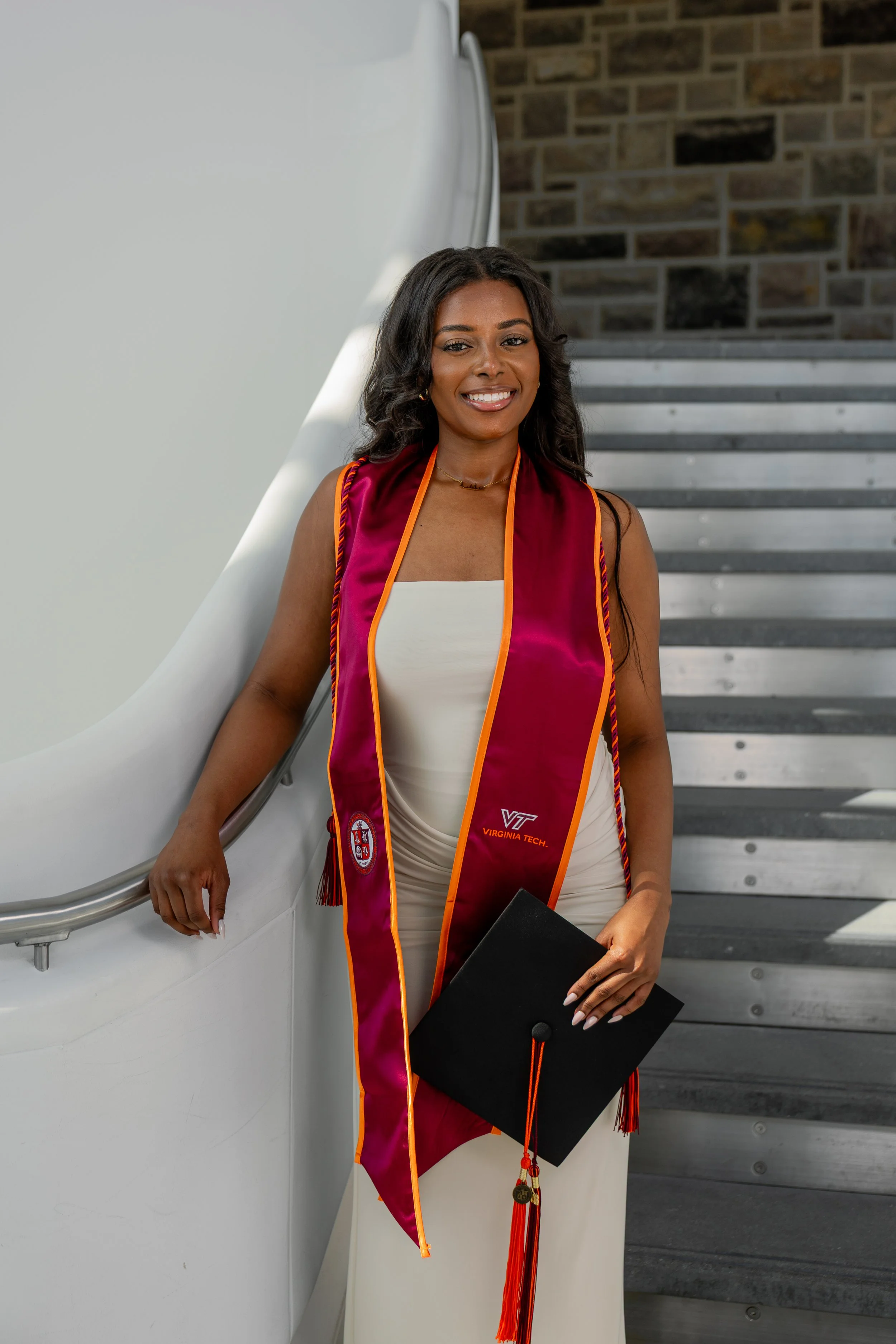 A woman in a graduation cap and gown standing on stairs, smiling, with a brick wall in the background.