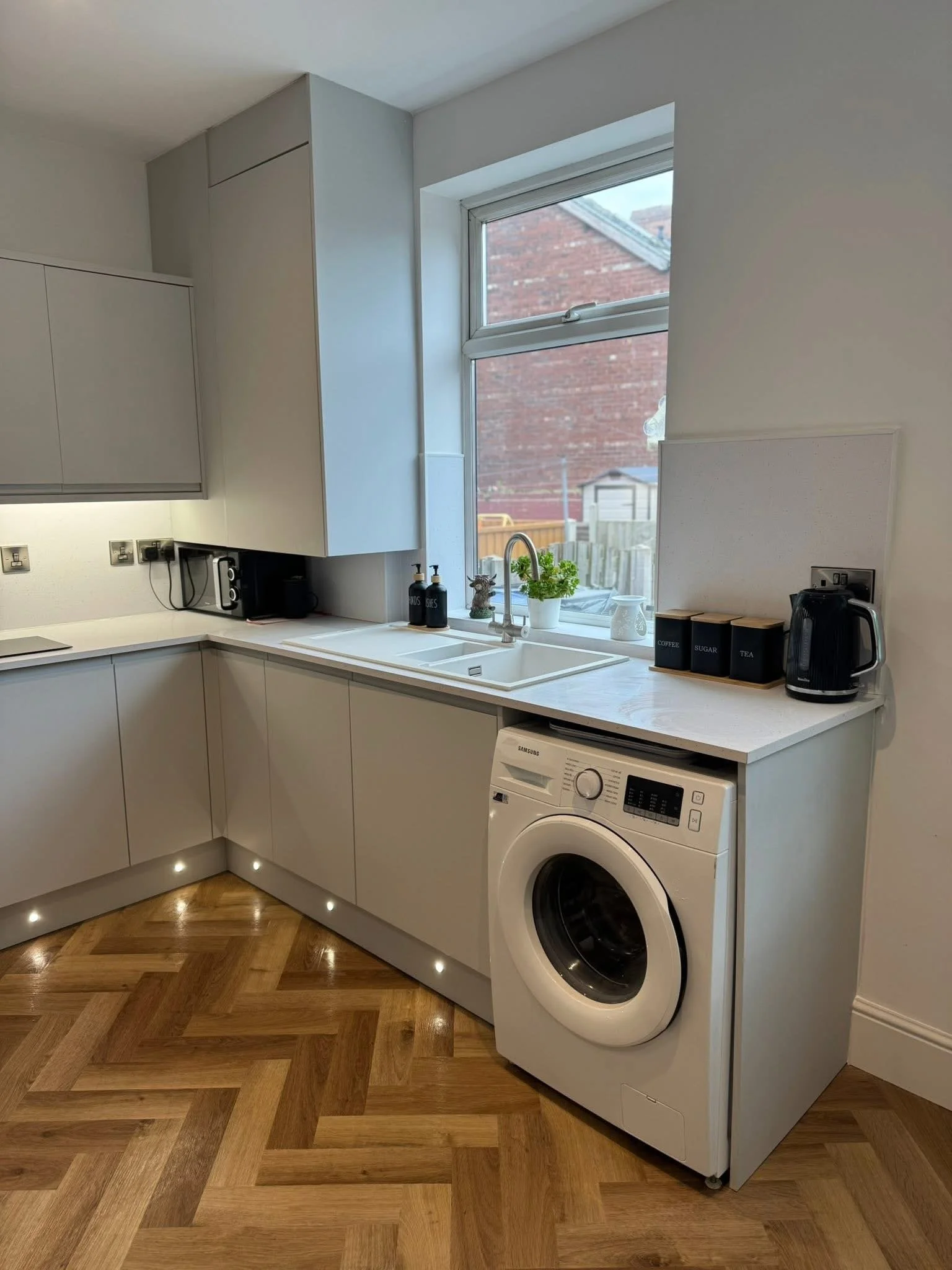 A modern kitchen with white cabinets, a large window above the sink, a washing machine under the counter, and various kitchen items including a plant, soap dispensers, and canisters labeled 'Coffee,' 'Sugar,' and 'Tea'.